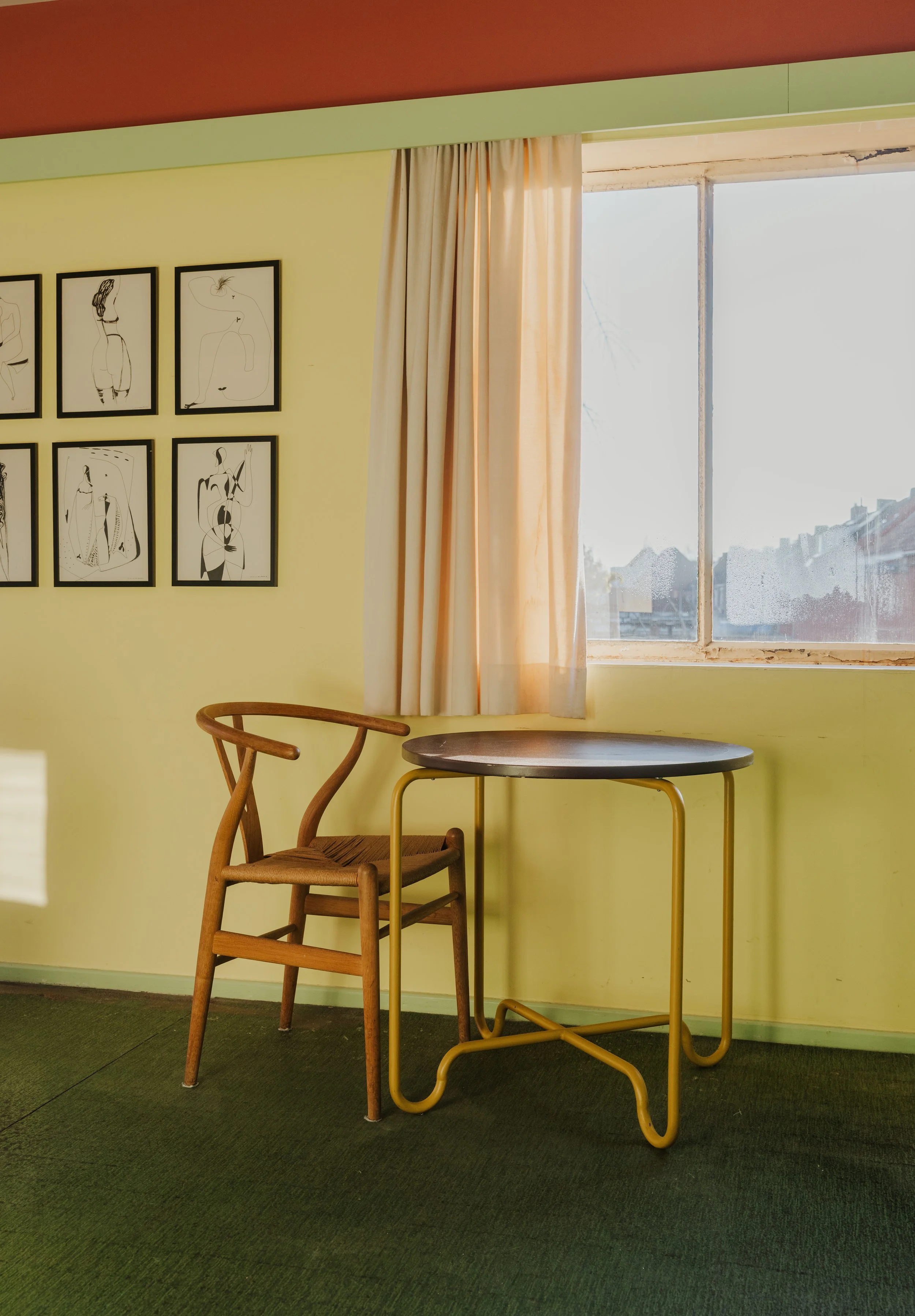 Interior of a room with yellow walls, a wooden chair, round table with yellow metal legs, framed abstract line art on the wall, window with cream curtains, and green carpet flooring.