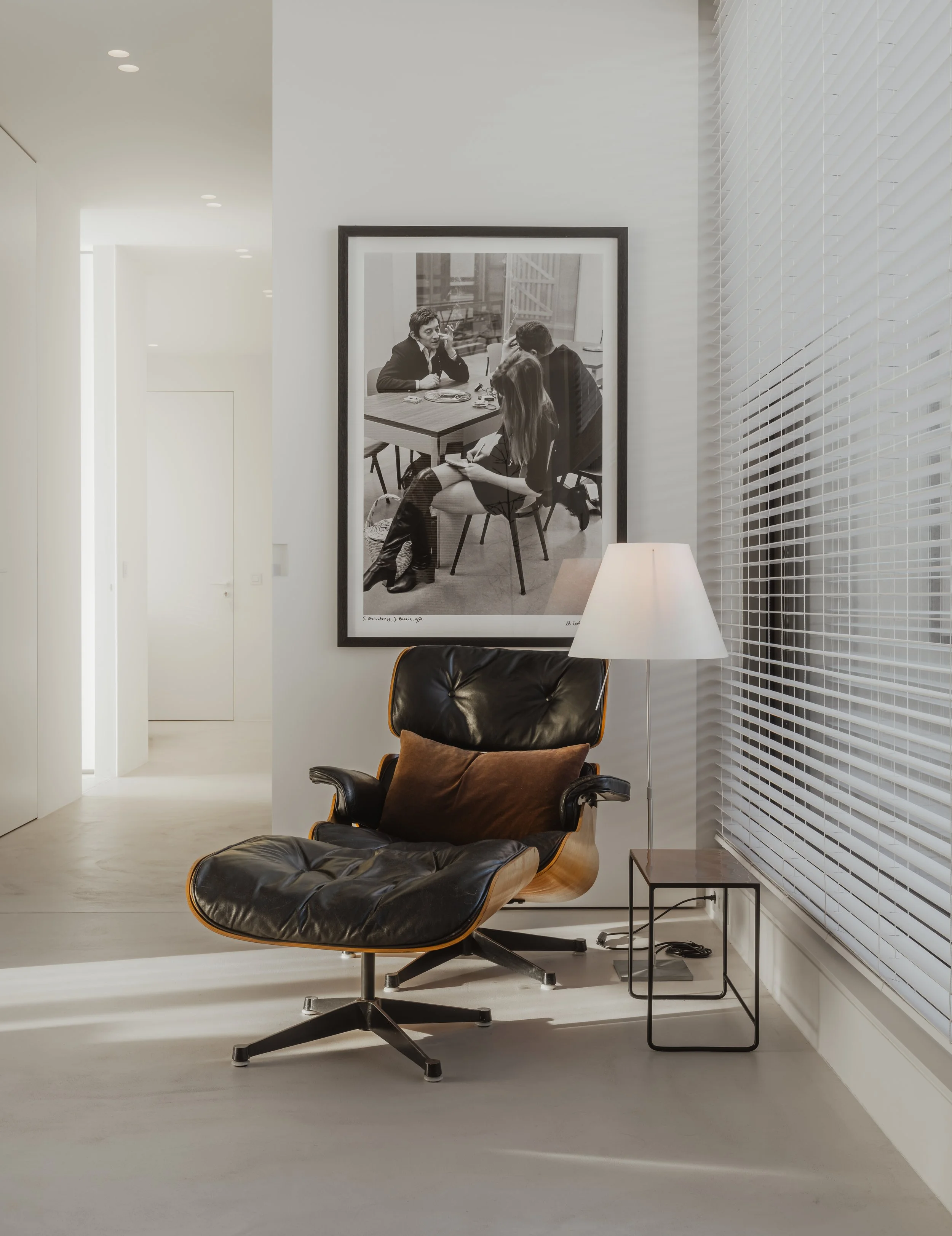 Modern living room with a black leather lounge chair, a side table with a lamp, a large framed black-and-white photograph on the wall, and white blinds on the window.