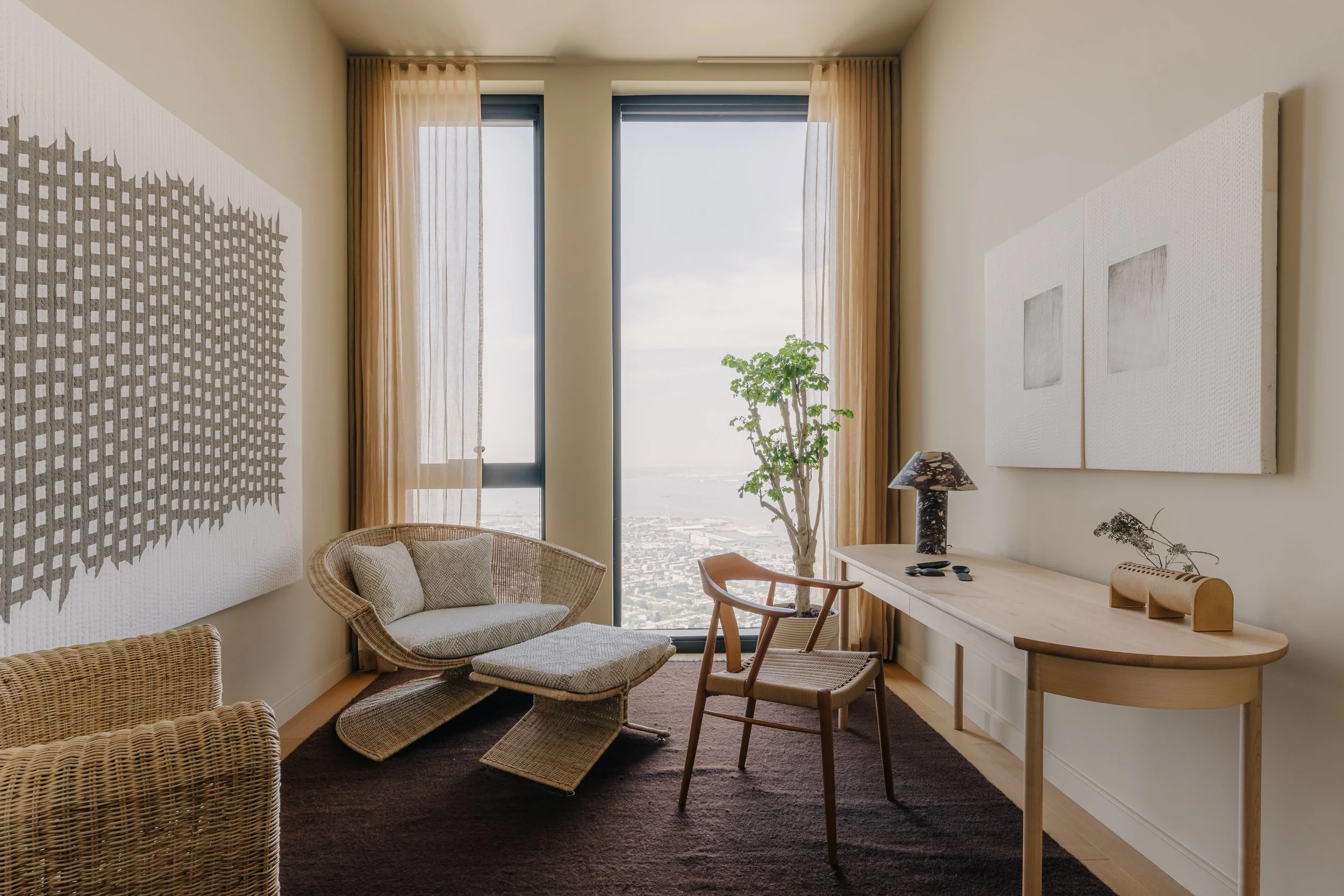 A modern living room with large windows, sheer curtains, a rattan armchair with a pillow, abstract wall art, a wooden table with decorative items, a potted tree, and city view outside.