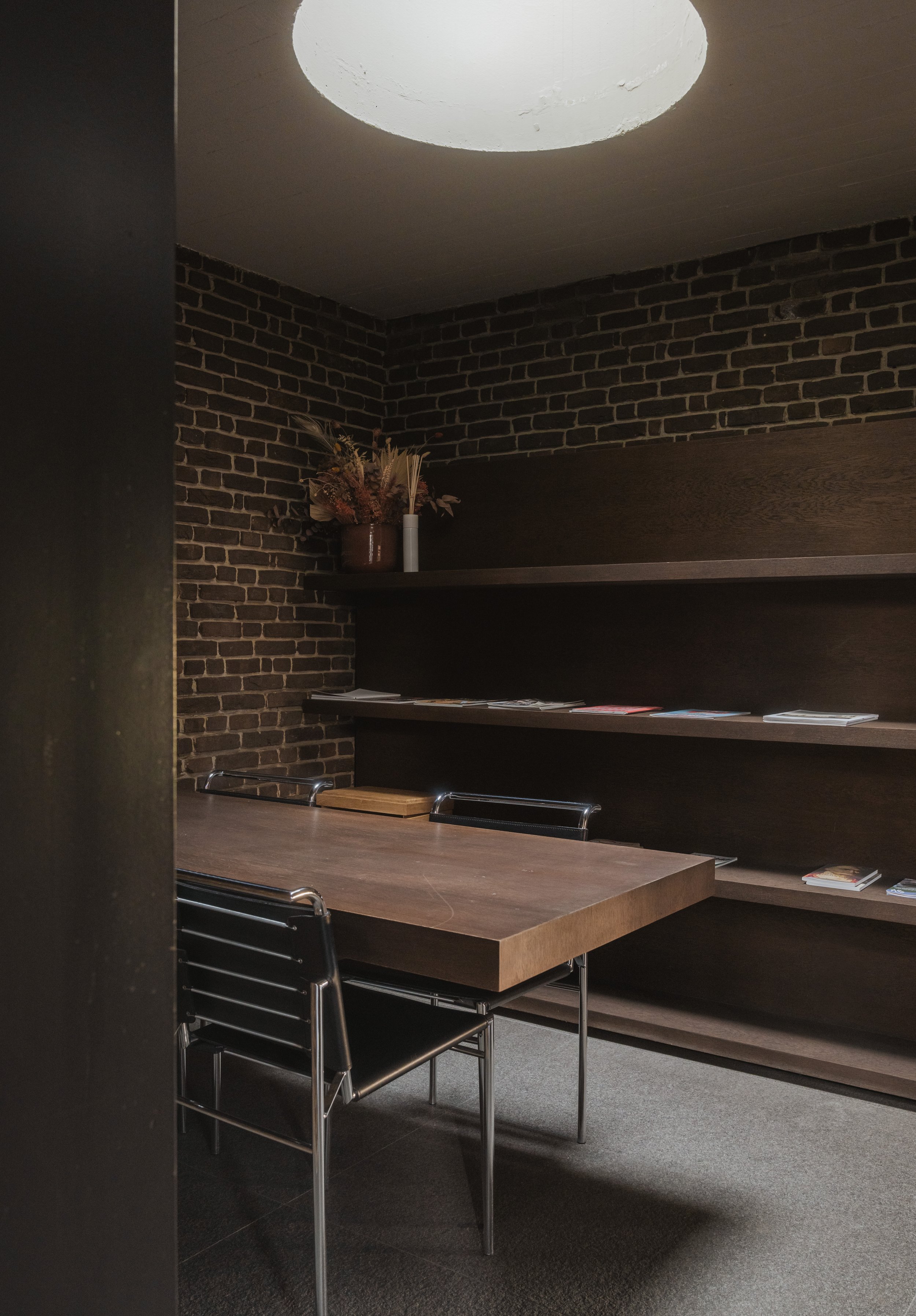Interior of a small meeting room with a wooden table and black chairs, dark brick wall with shelves holding magazines, and a vase with dried flowers on top, illuminated by a circular ceiling light.
