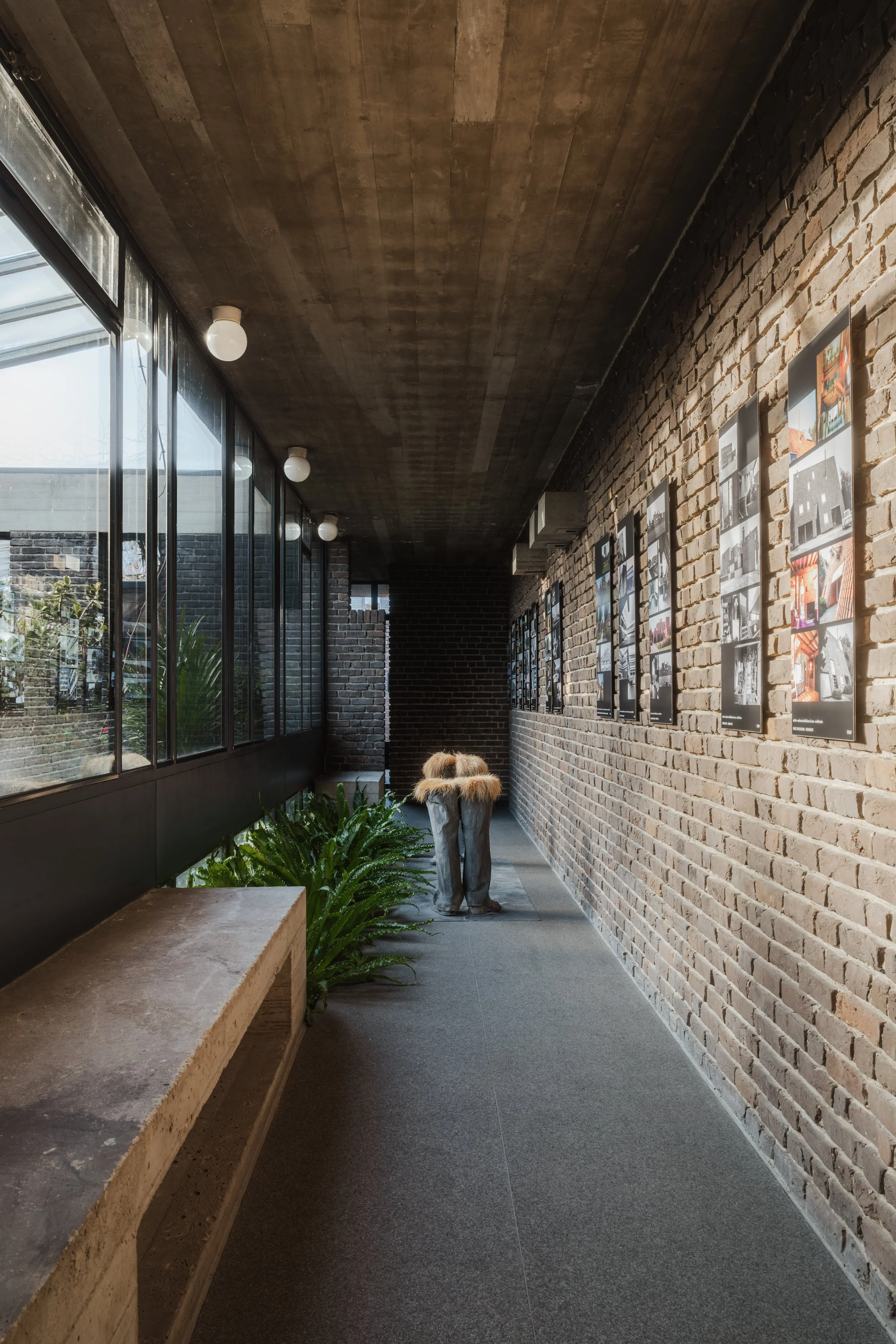 Indoor hallway with brick walls, large windows, and framed photographs on the right wall. There is a bench and some potted plants along the left side. The ceiling has exposed wood and white globe lights.