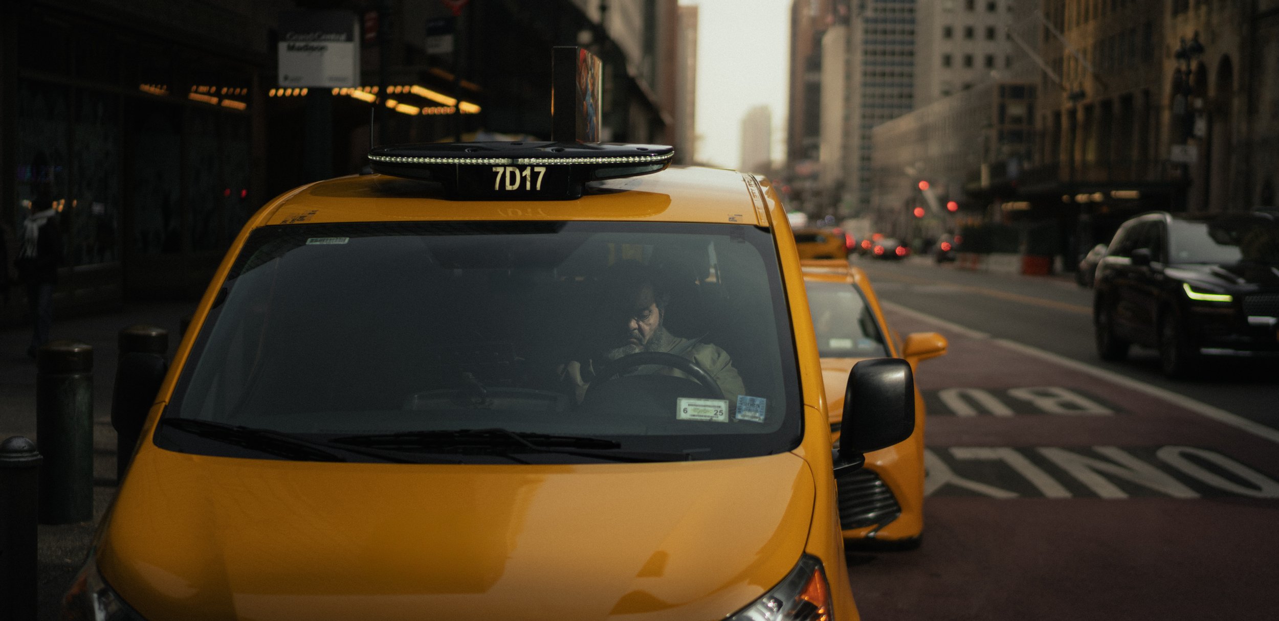 A yellow taxi cab is stopped on the city street, with a person sitting inside behind the wheel. Other yellow taxis and cars are visible in the background, and tall buildings line the street at sunset or dusk.