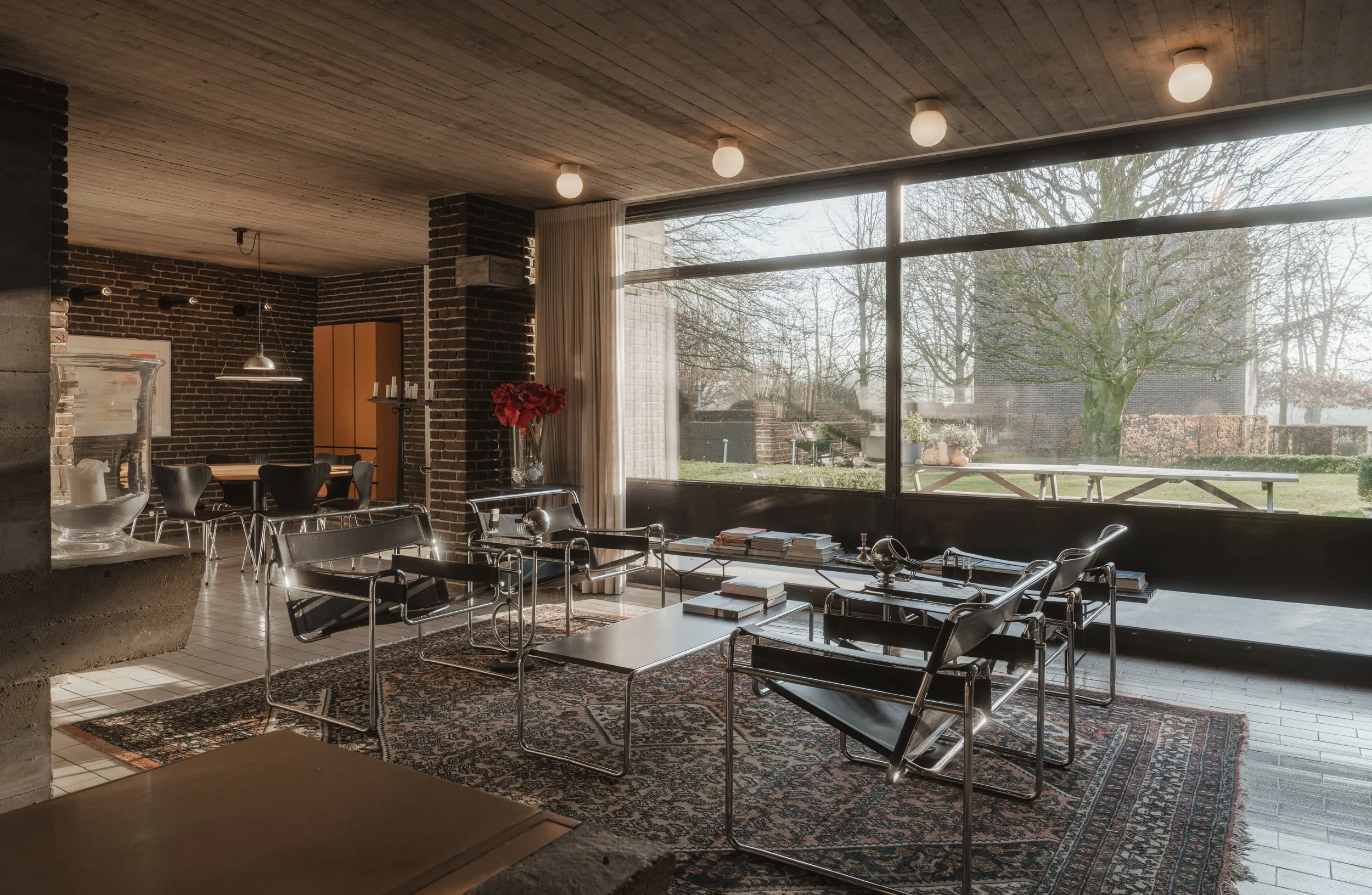 Living room with large window overlooking a backyard, modern black chairs, glass tables with books, brick walls, and a patterned rug.