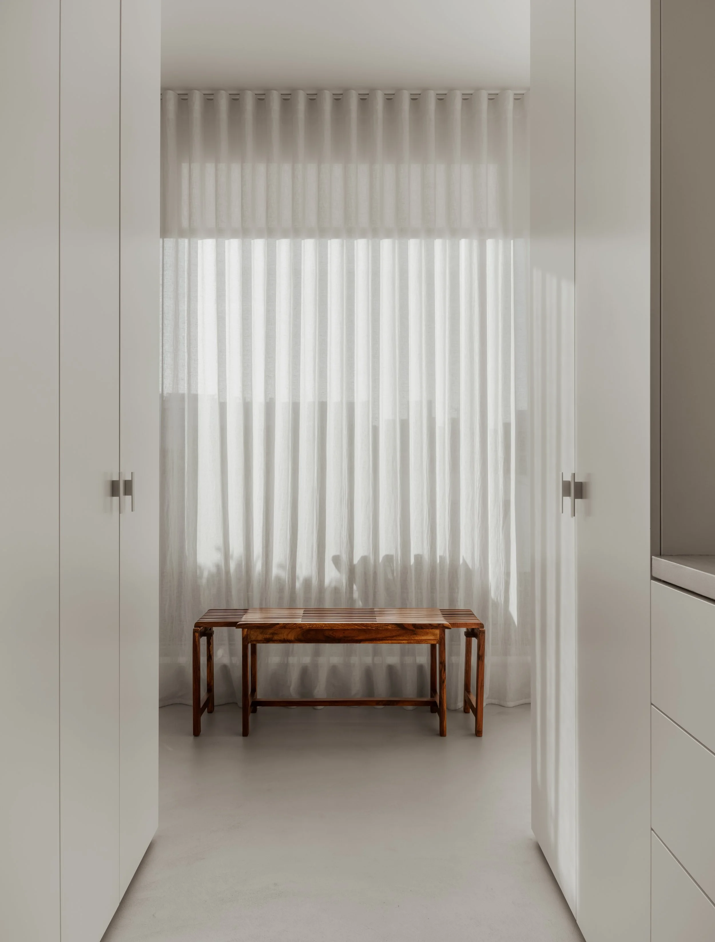 A simple room with white cabinetry framing a large window with sheer white curtains, a wooden bench beneath the window, and soft natural light.