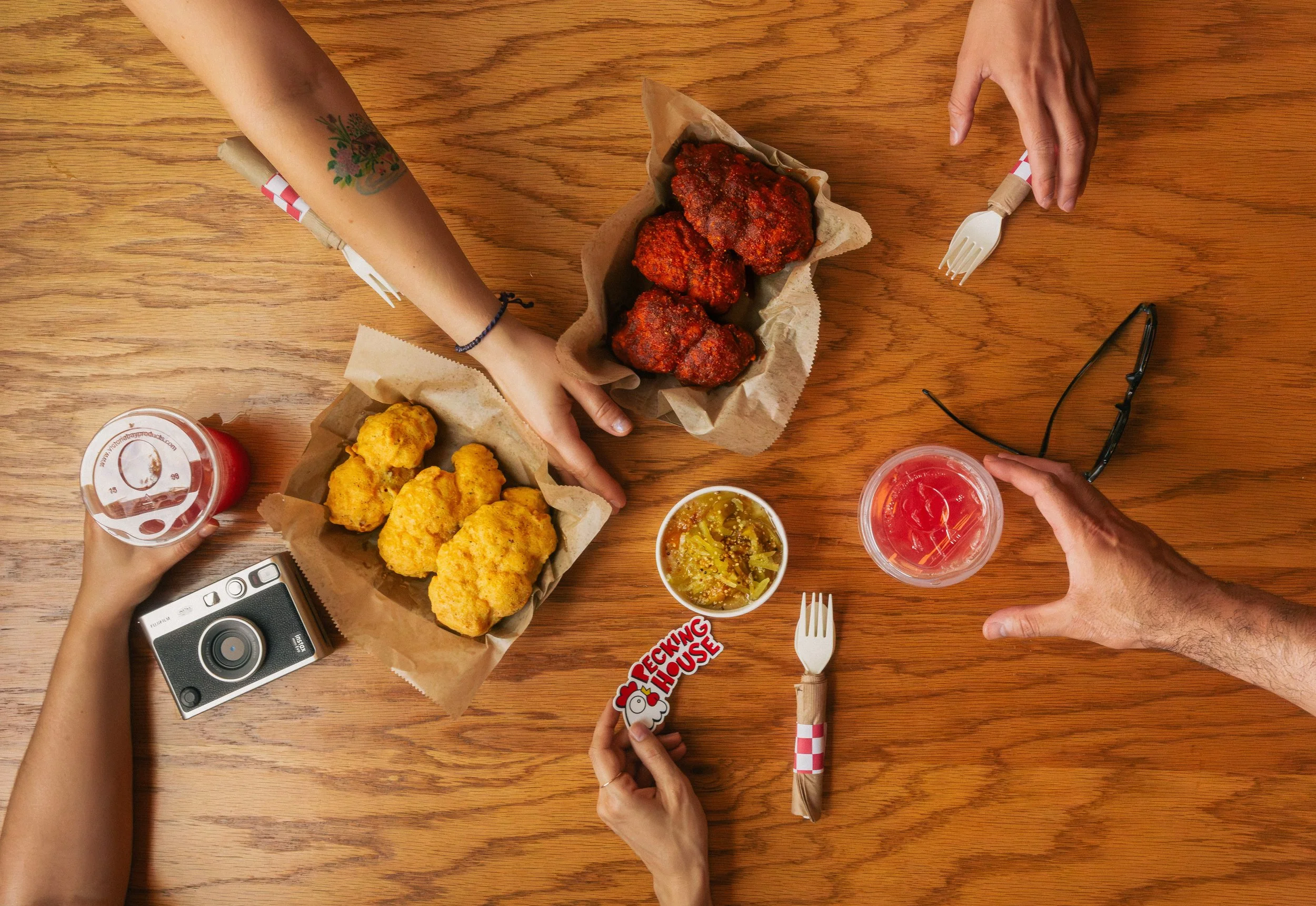 Overhead view of four people sharing a meal of fried chicken on a wooden table. The table has a plastic cup with a purple drink, a cup with pickles, a camera, and a hand holding a sticker that says 'Fucking House'.