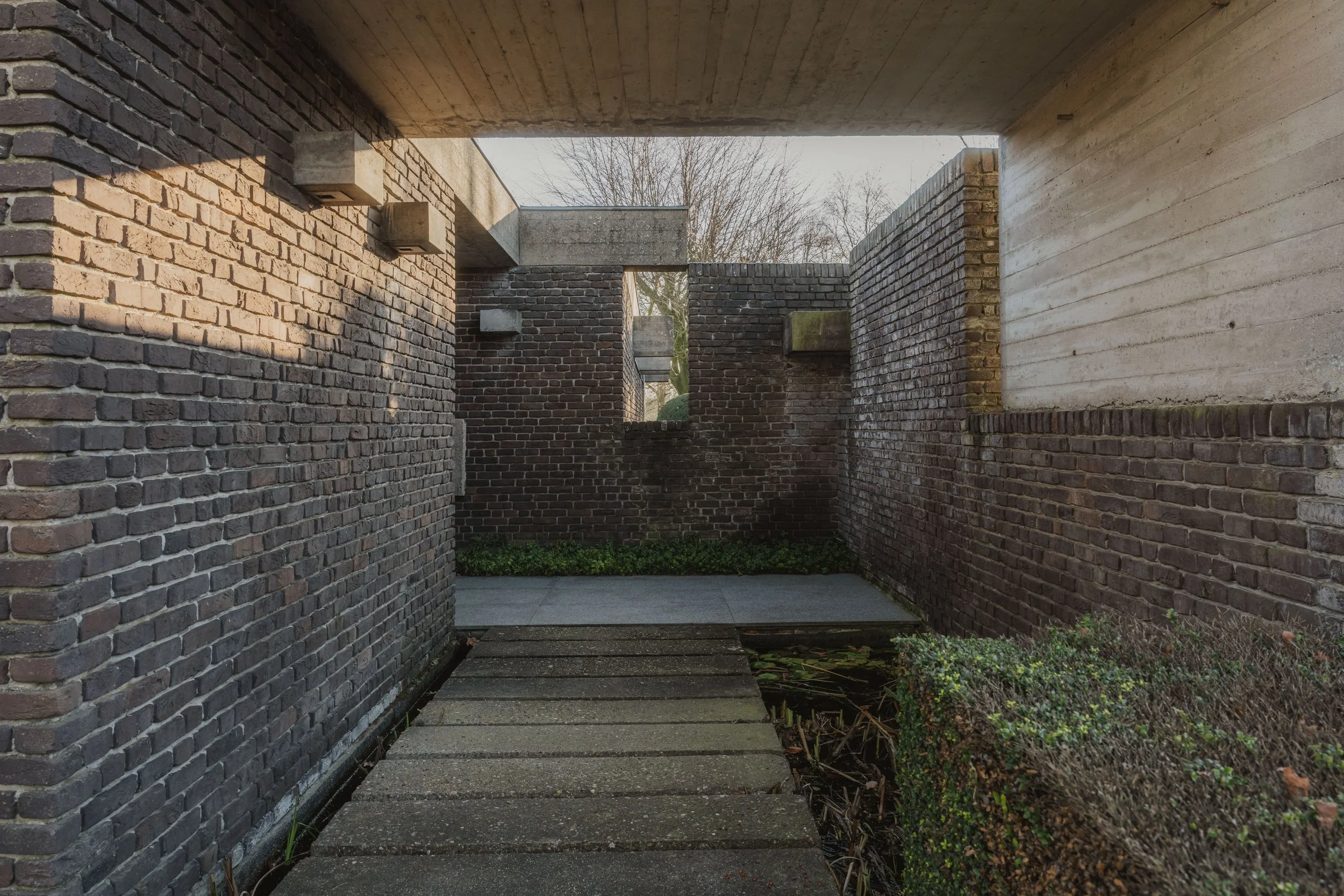 A narrow passage with brick walls and concrete ceiling, leading to a small courtyard with plants and a head of trees visible.