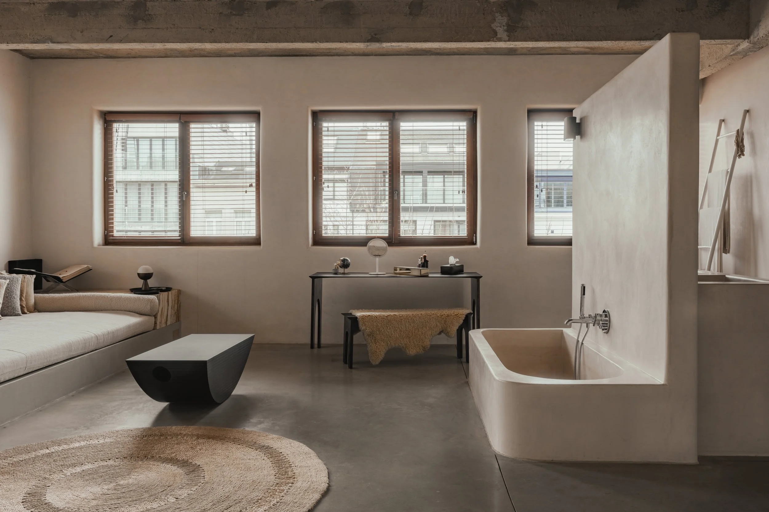 Modern minimalist living room with three large windows, a beige sofa on the left, a black coffee table, a small desk with decorative items in the center, and a bathtub on the right, all with neutral tones and simple decor.