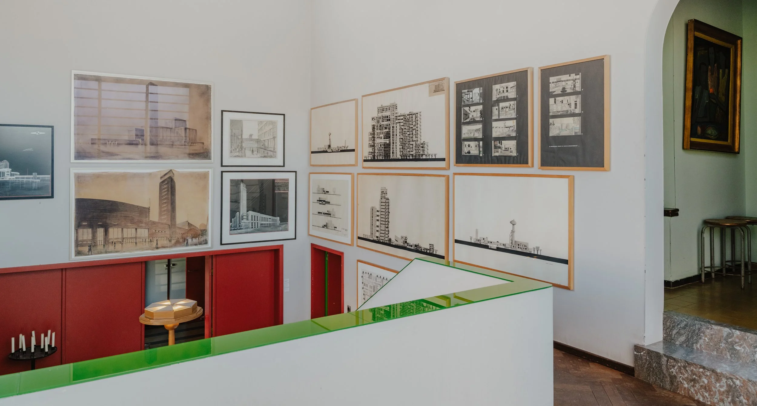 Gallery with framed architectural drawings and photographs on white wall, a red storage cabinet, an art installation with candles, and a separate room with a painting on a green wall.