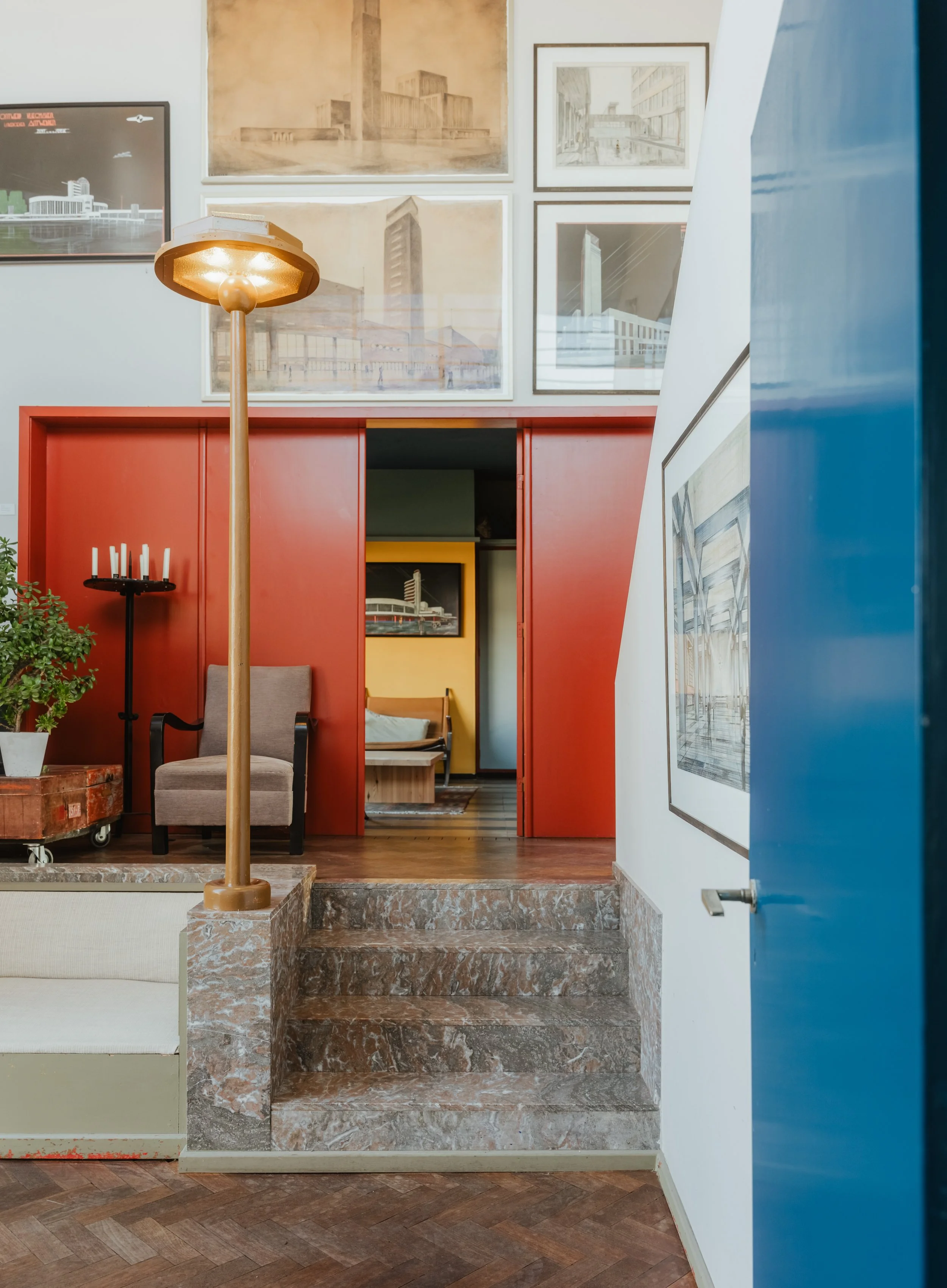 Interior of an art gallery or museum with stairs leading to an upper level, framed sketches and architectural drawings on the walls, a chair, a plant, and a lit floor lamp.