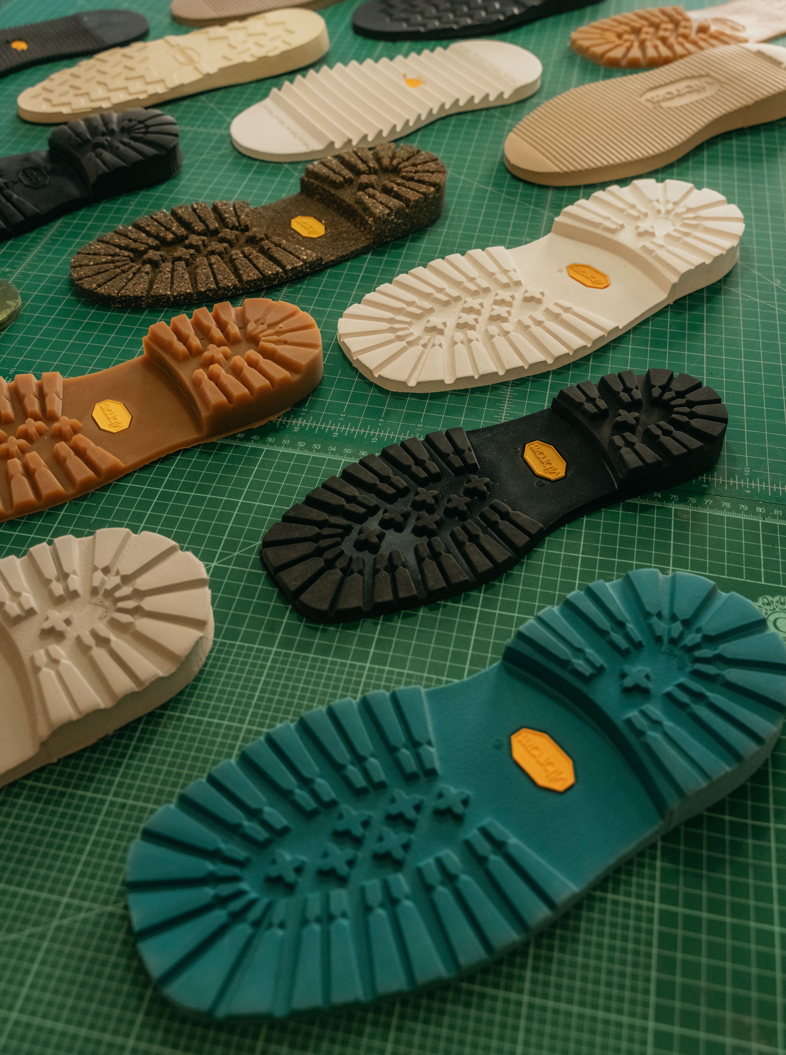 Multiple colorful shoe soles laid out on a green cutting mat, showcasing various tread patterns and colors including black, white, tan, blue, and gray.