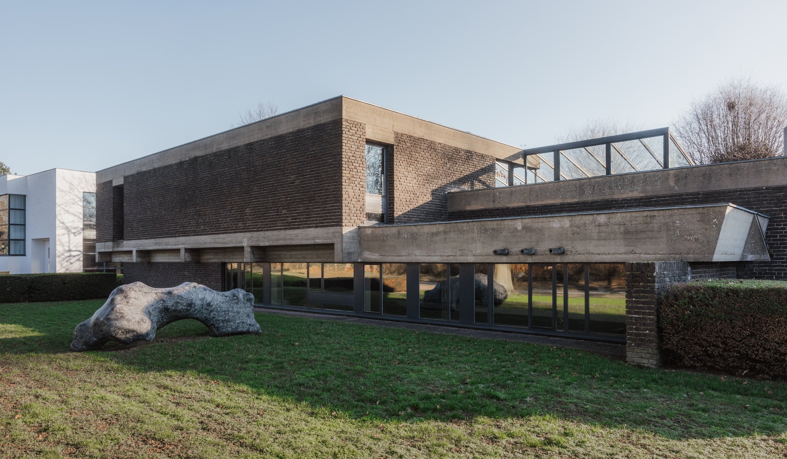 Modern building with brick and concrete exterior, large windows, and a glass-railed rooftop terrace. There is a large rock and neatly trimmed grass in the foreground.