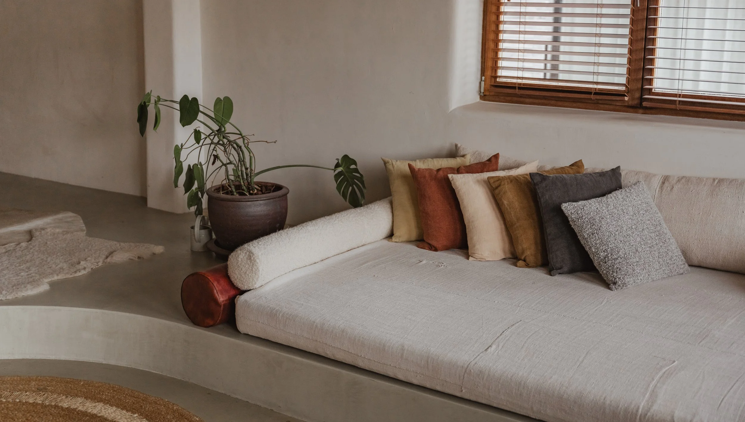 Living room with a built-in beige sofa, colorful throw pillows, a potted Monstera plant, and wooden window blinds.