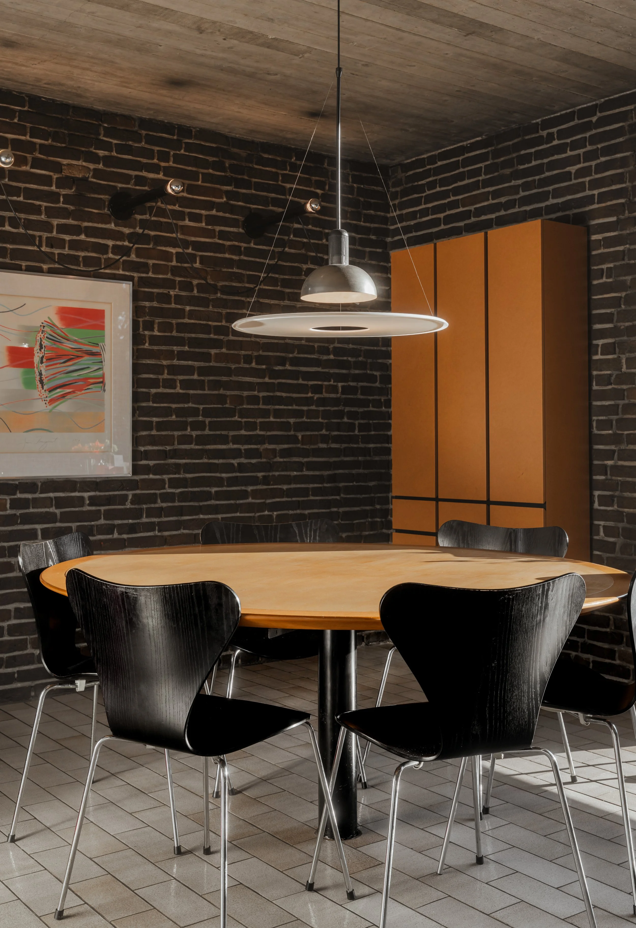 Modern dining room with a round wooden table surrounded by six black chairs, a metallic pendant light above, dark brick walls, a colorful abstract painting, and a tall orange cabinet.
