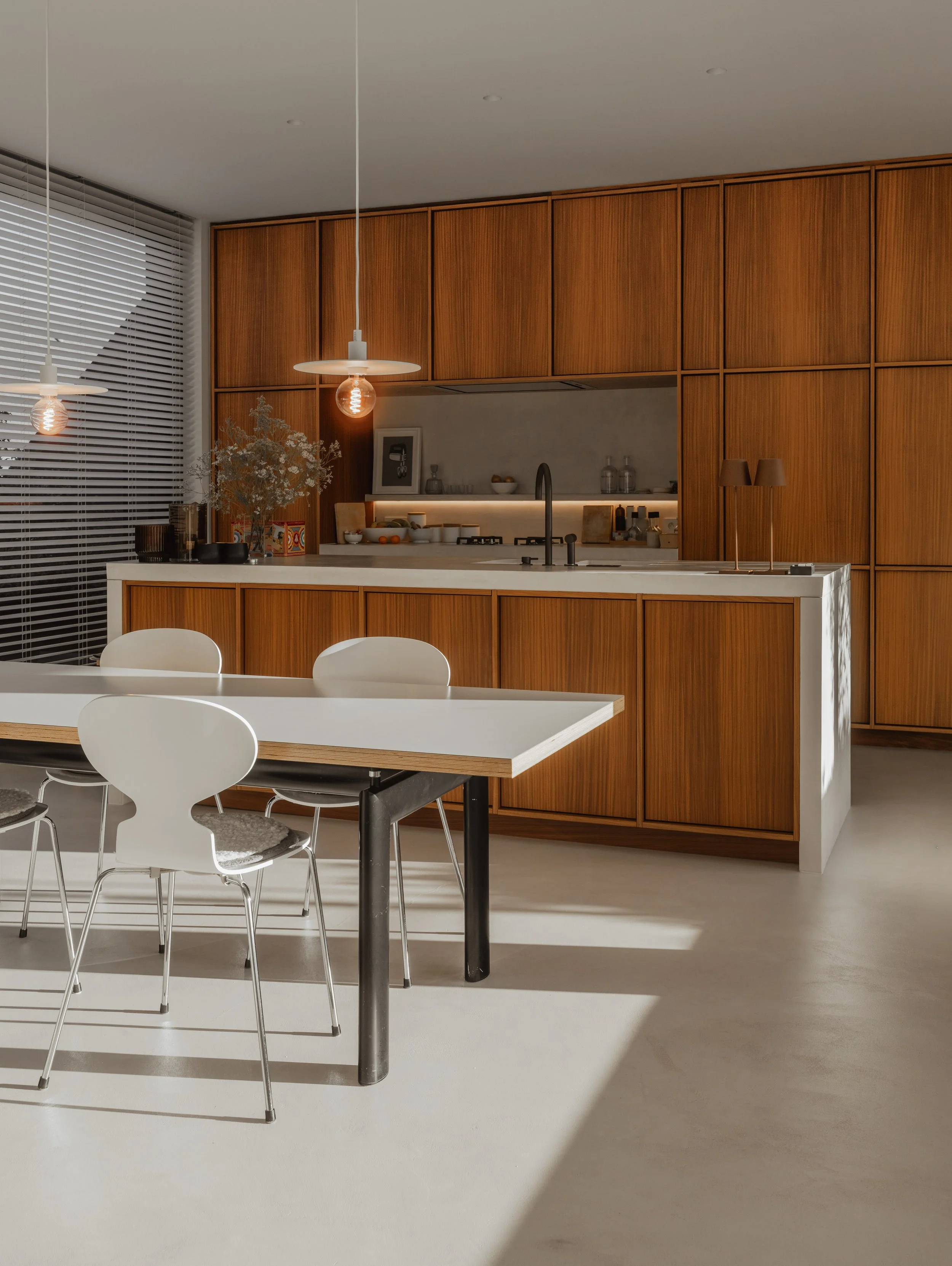 Modern kitchen with wooden cabinets, a white island, and a dining table with white chairs. Two hanging pendant lights above the island, and sunlight casting shadows on the floor.