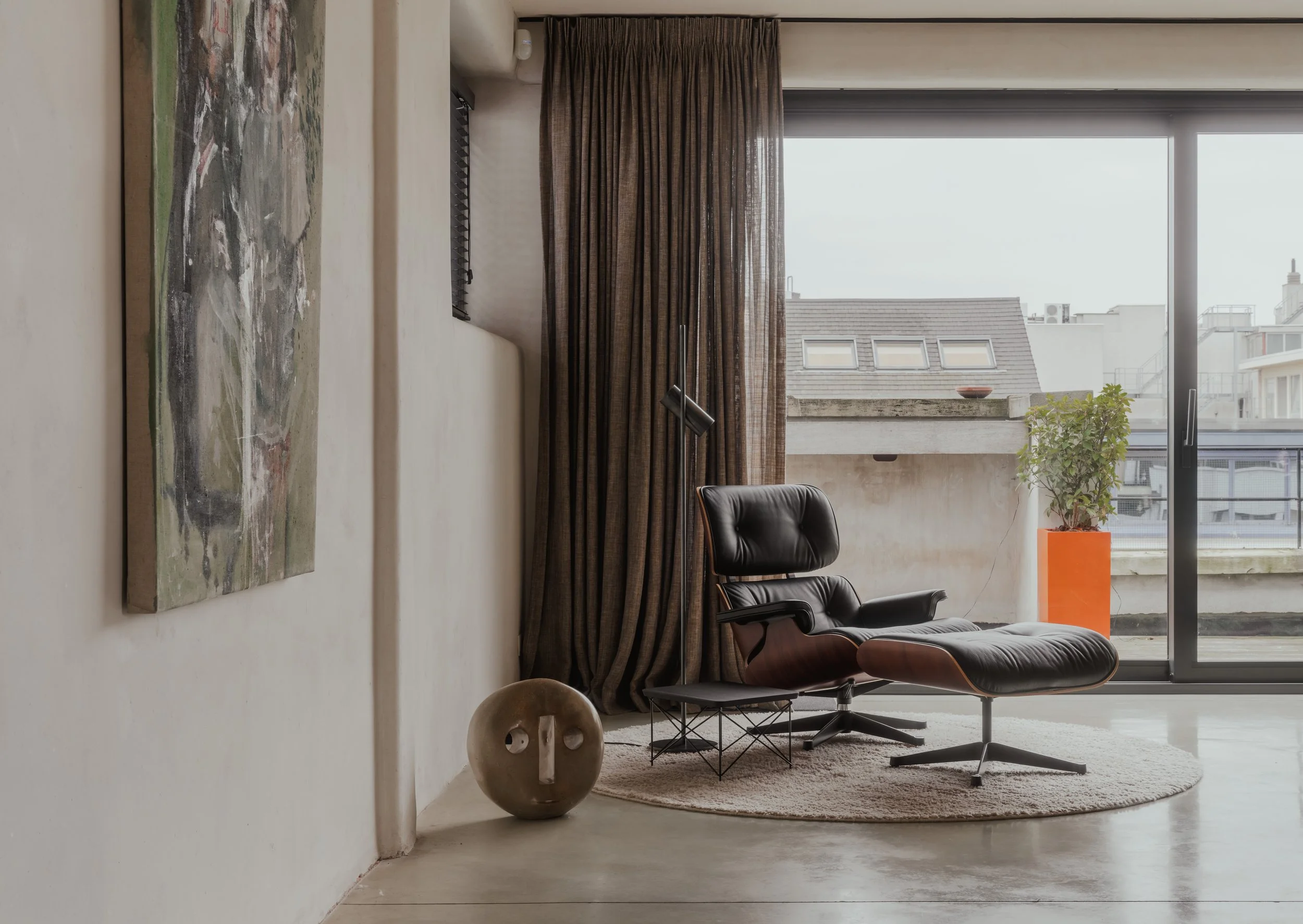 Modern living room with a black leather lounge chair and ottoman on a round beige rug, floor-to-ceiling window with brown curtains, potted plant in an orange pot outside, abstract artwork on the wall, ceramic face sculpture on the floor, and city bui