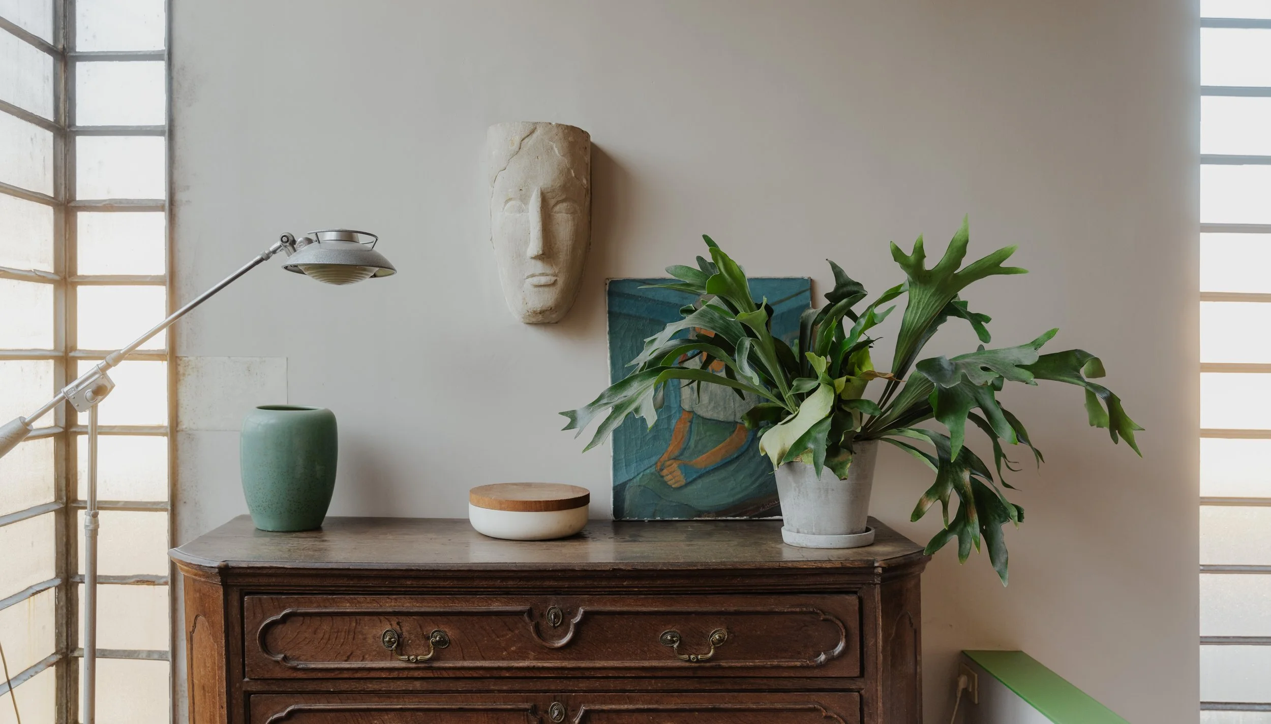 Decorative shelf with a green vase, a white and wood container, a painting, and a large potted plant on a wooden cabinet, with a stone face sculpture on the wall above