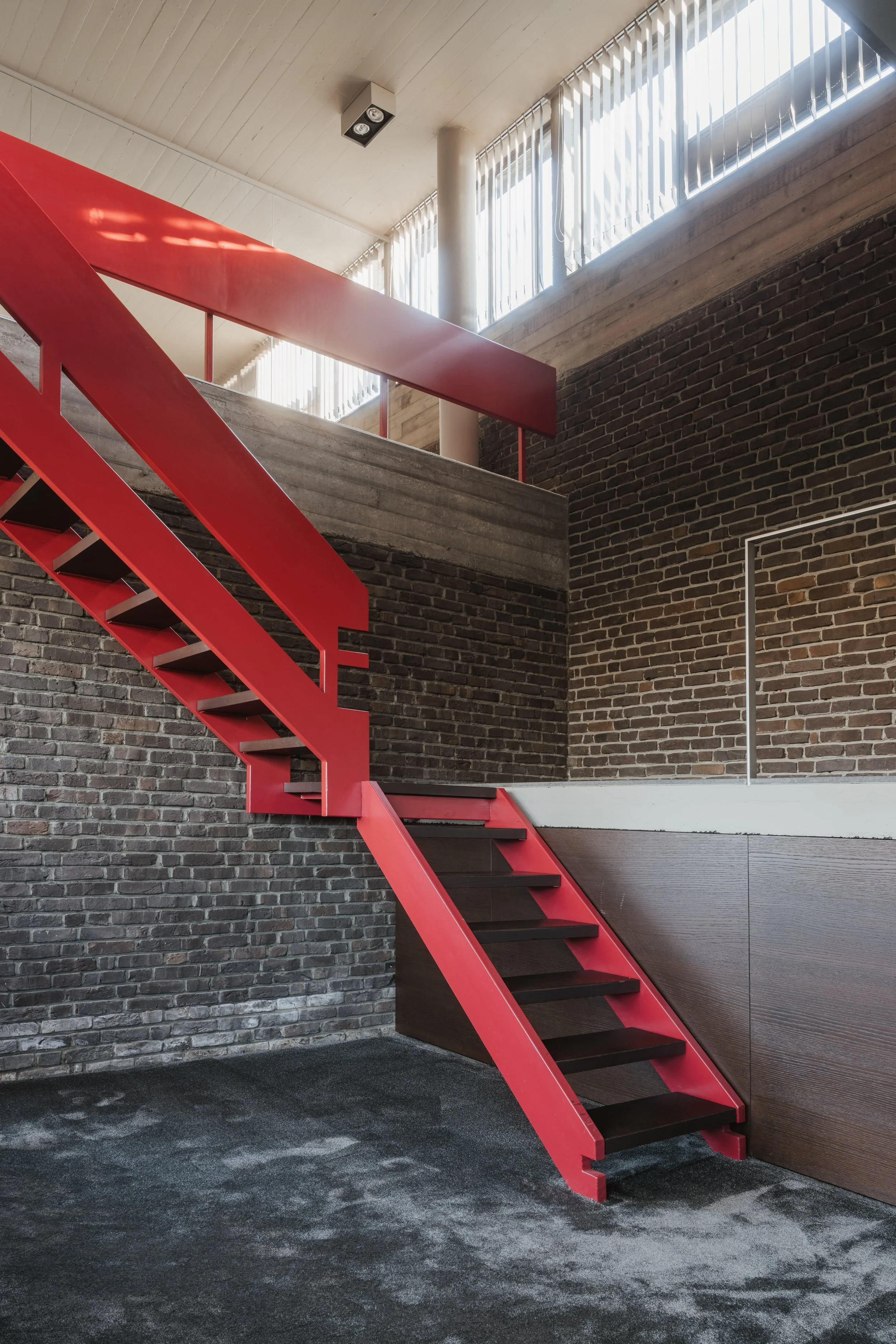 Interior of a modern building featuring a red staircase, brick wall, and large windows with vertical blinds.