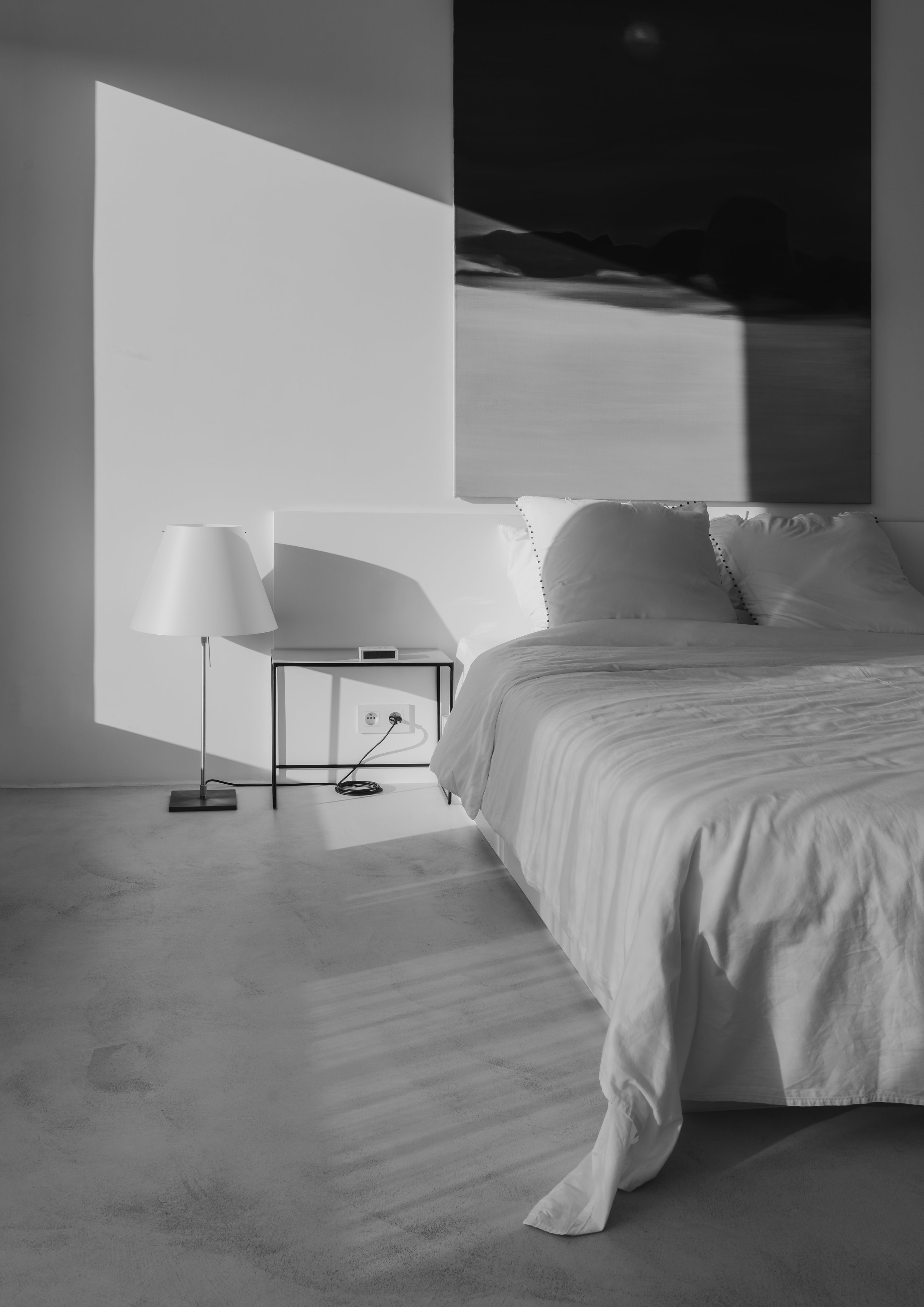 A minimalist bedroom with a white bed, white pillows, a bedside table, a lamp, and a large landscape painting on the wall. Sunlight creates shadows on the wall and floor.