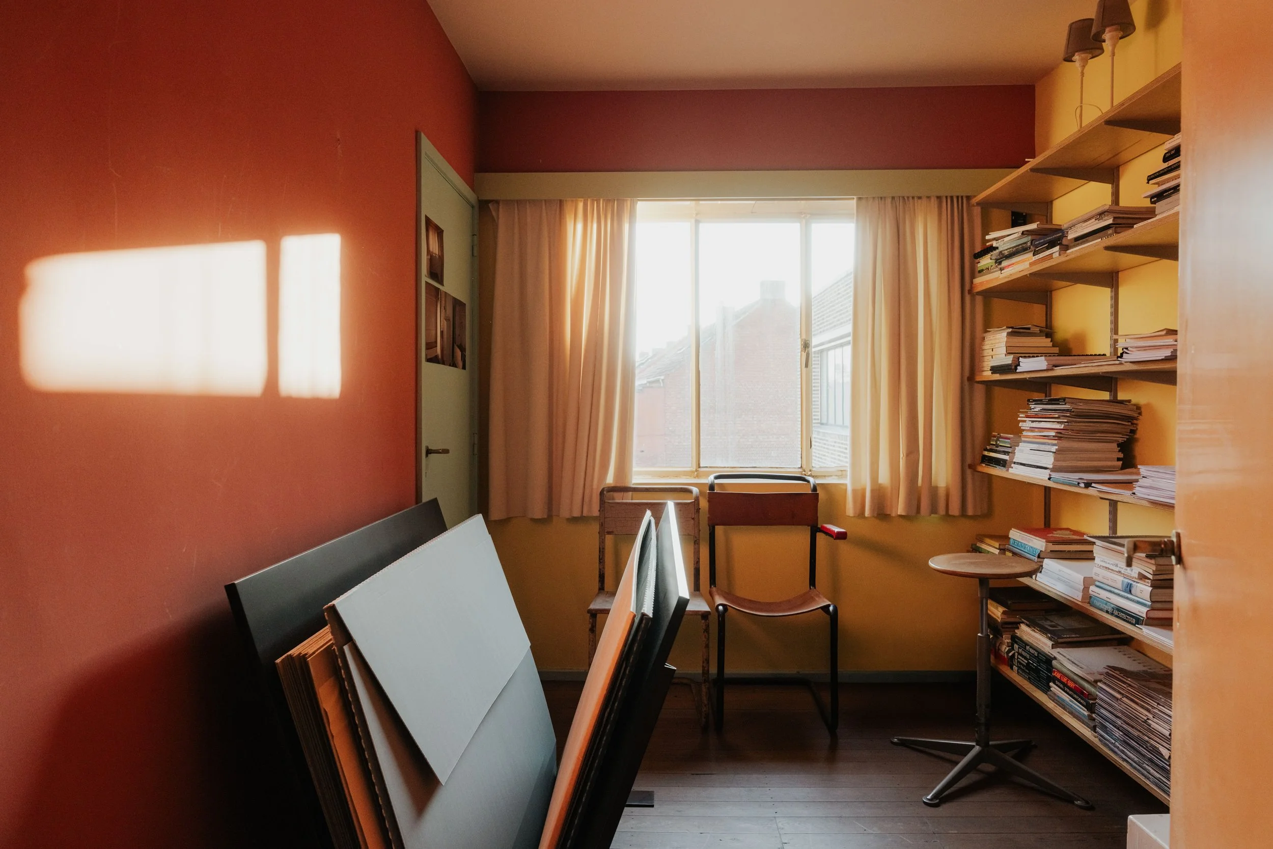 A small room with a window letting in sunlight, beige curtains, a bookshelf filled with papers and books, and two chairs by the window.