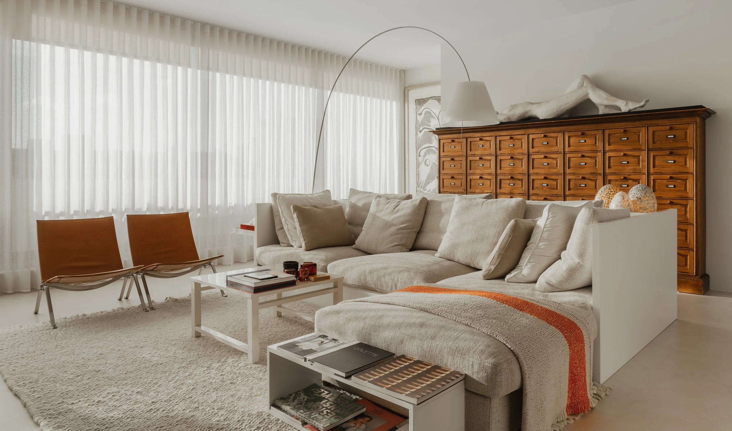 Living room with white sofa, beige cushions, white coffee table, two brown chairs, large window with sheer white curtains, wooden cabinet with multiple drawers, and decorative lamps.