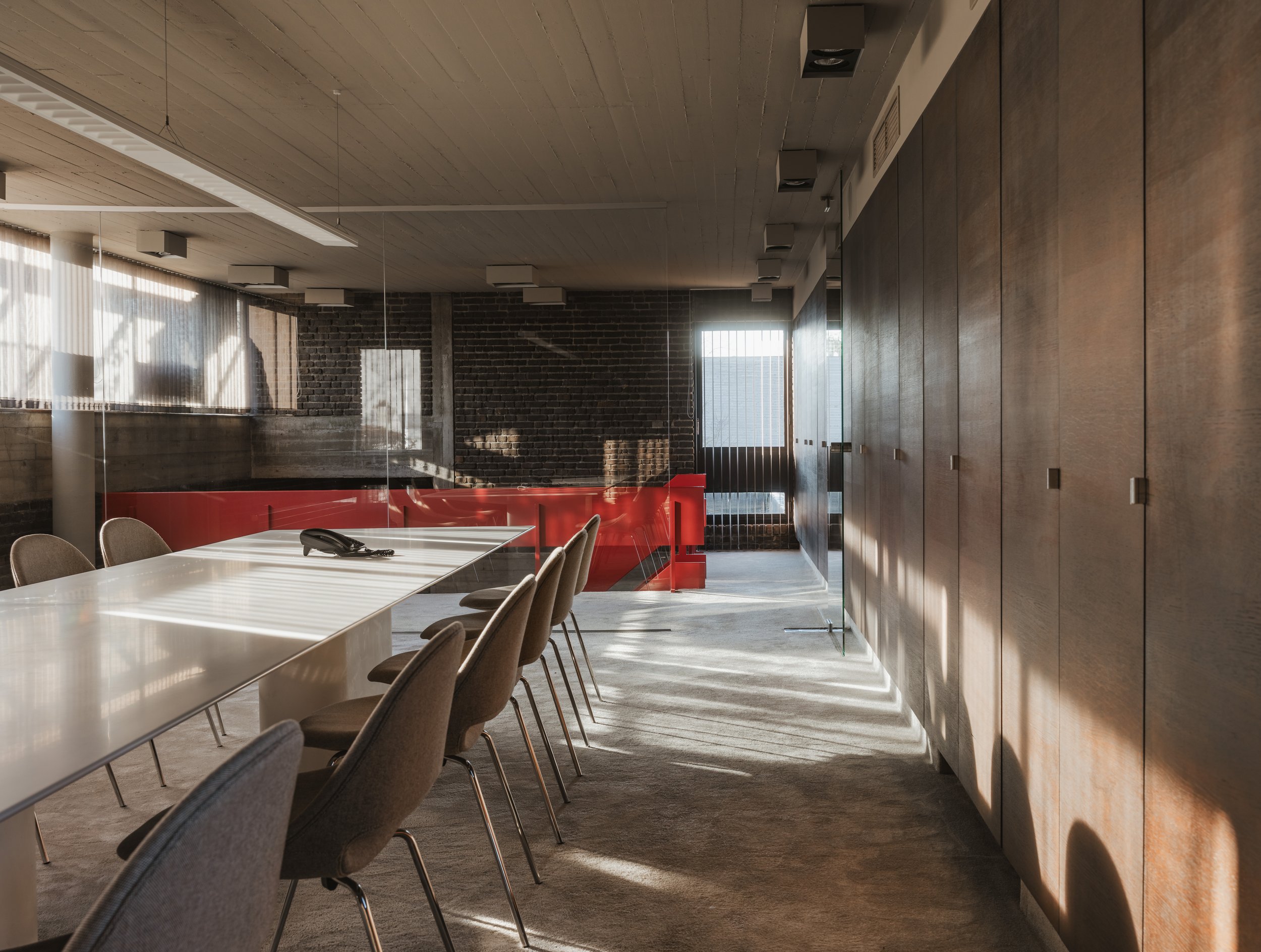 Empty conference room with a long white table, beige chairs, a black telephone, and wooden cabinets on the right side. Large windows with sheer curtains let in sunlight, creating shadows on the carpeted floor.