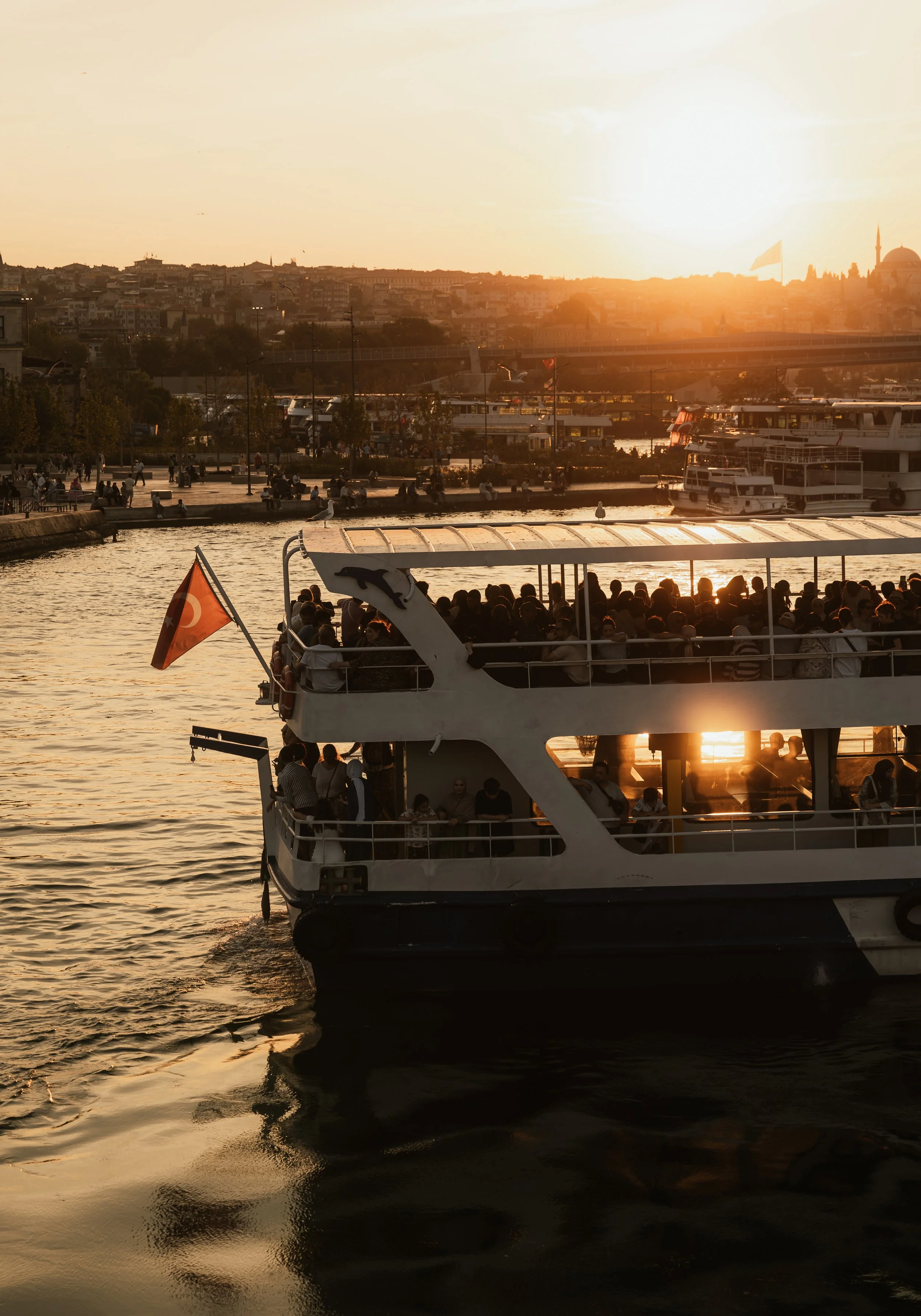 A ferry boat filled with people sailing on the water during sunset with a city skyline and other boats in the background.