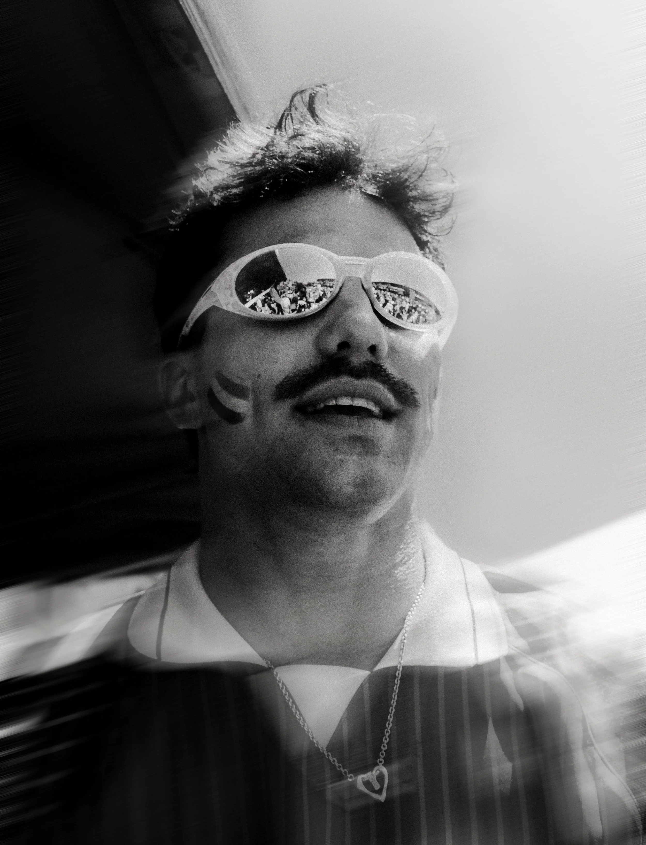 A black-and-white selfie of a man with curly hair, wearing sunglasses with reflective lenses, a white T-shirt, and a chain necklace with a heart-shaped pendant. The reflection in his sunglasses shows a crowd or group of people and part of a building.