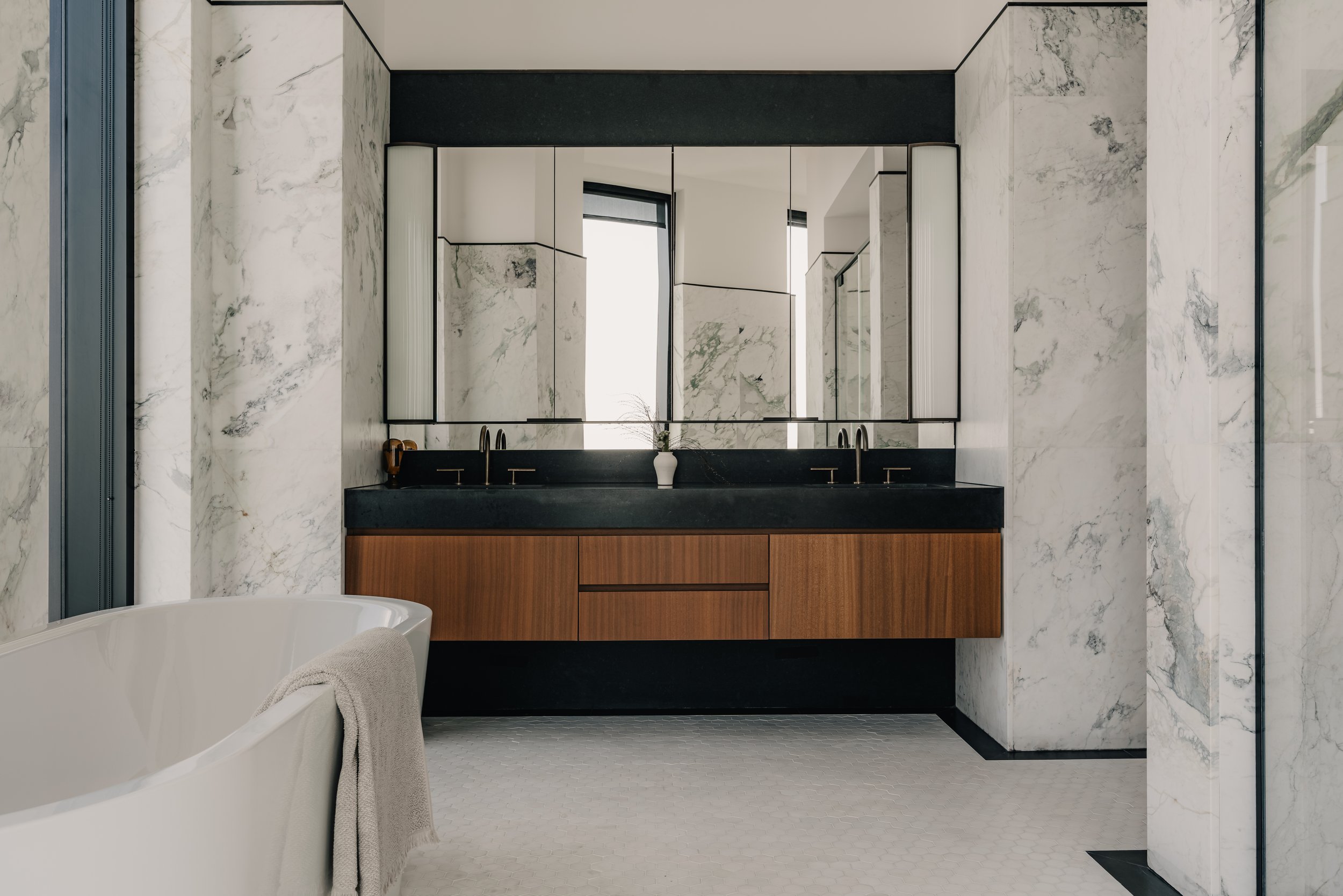 Modern bathroom with marble walls, a large mirror, wooden vanity with black countertop and two sinks, a white bathtub with a towel, and a window with black blinds.
