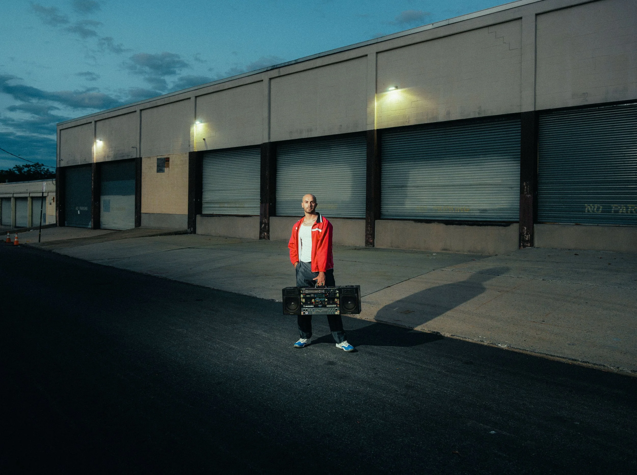 A young man in a red jacket standing on an empty street at dusk, holding a large boombox, with a warehouse building in the background.