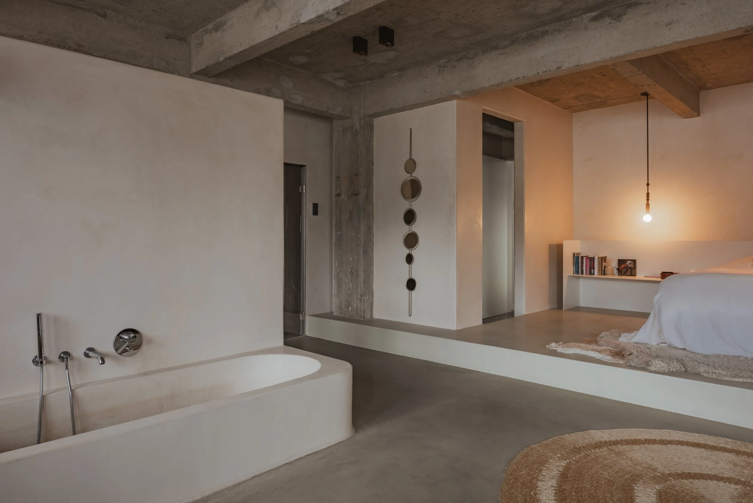 Interior view of a modern, minimalist bedroom with concrete and white walls. Visible are a built-in bathtub, a hanging light bulb, a bed with white bedding, a rug, and a shelf with books.