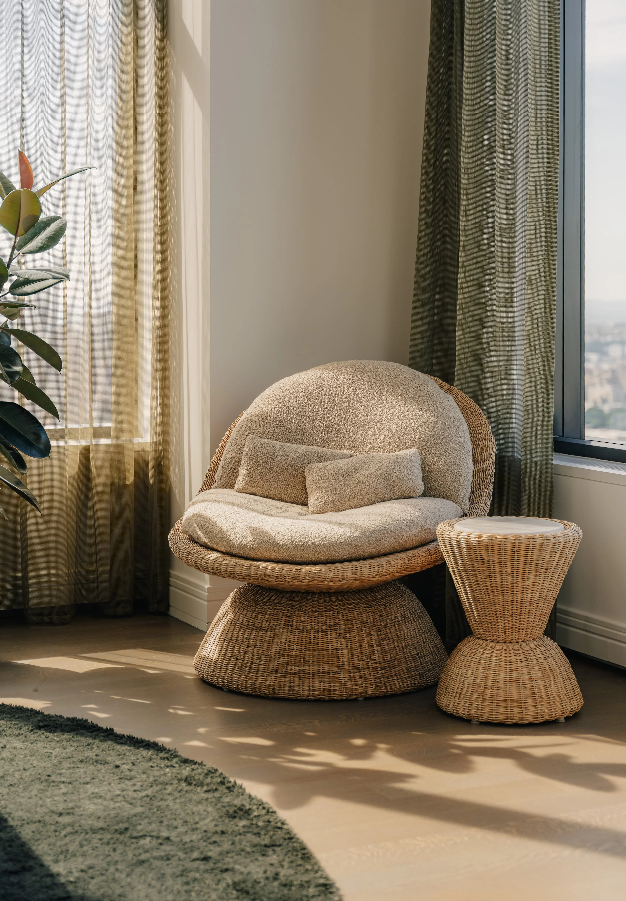 Sunlit corner of a modern living space with a wicker chair, a matching side table, plush beige cushions, a tall green houseplant, sheer curtains, and a large window showing a cityscape.