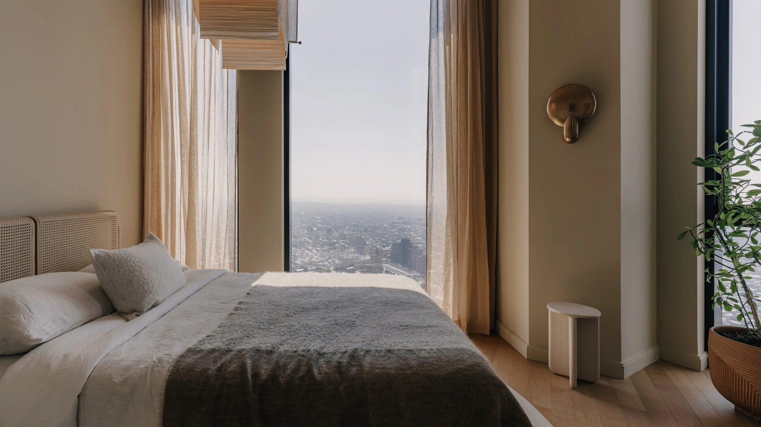Modern bedroom with a large window overlooking a cityscape, light-colored curtains, a bed with white bedding and pillows, a small white stool, a potted plant, and wall-mounted decor.