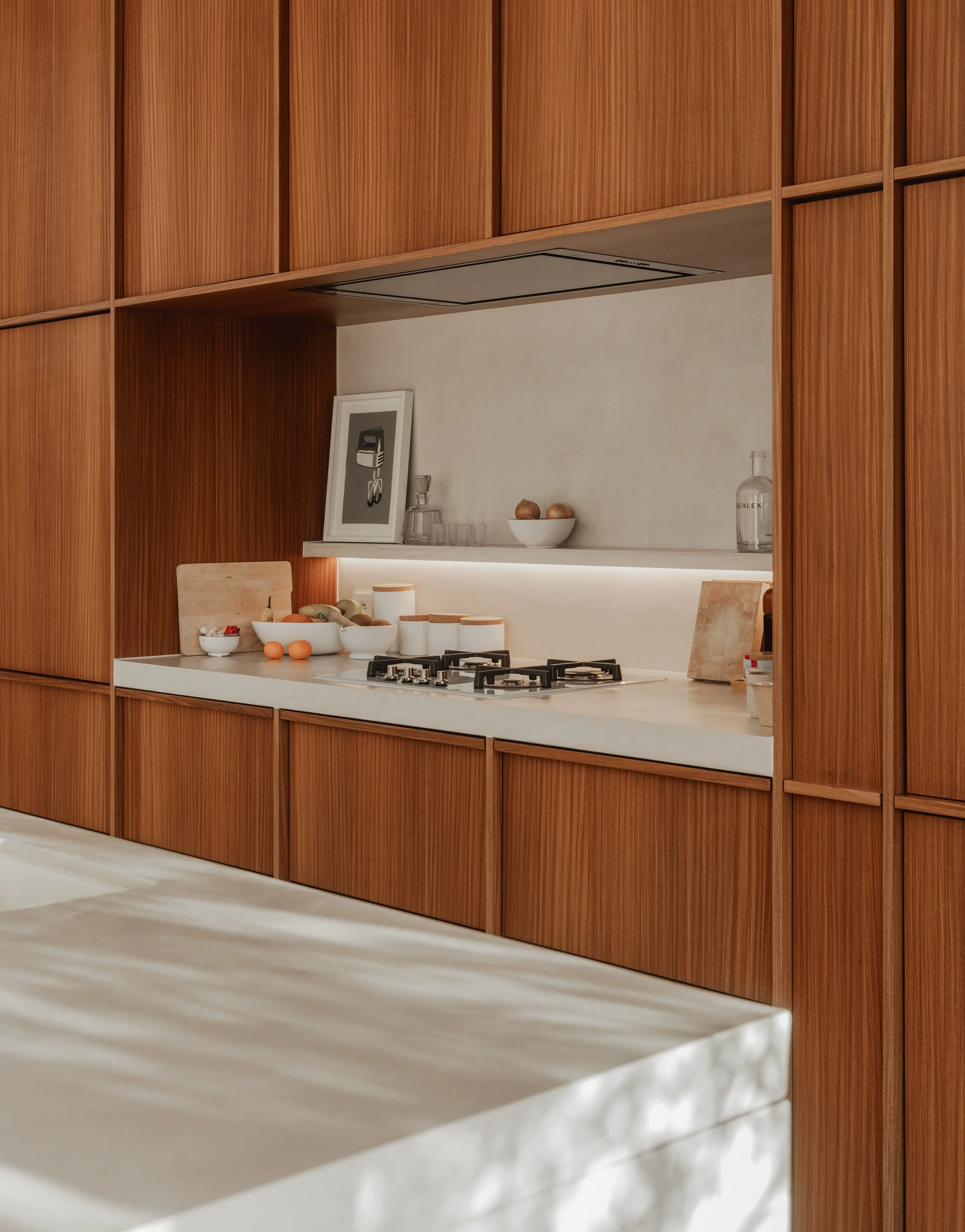 Modern kitchen with white countertop, wooden cabinets, and minimal decor including bowls of fruit and a framed artwork.