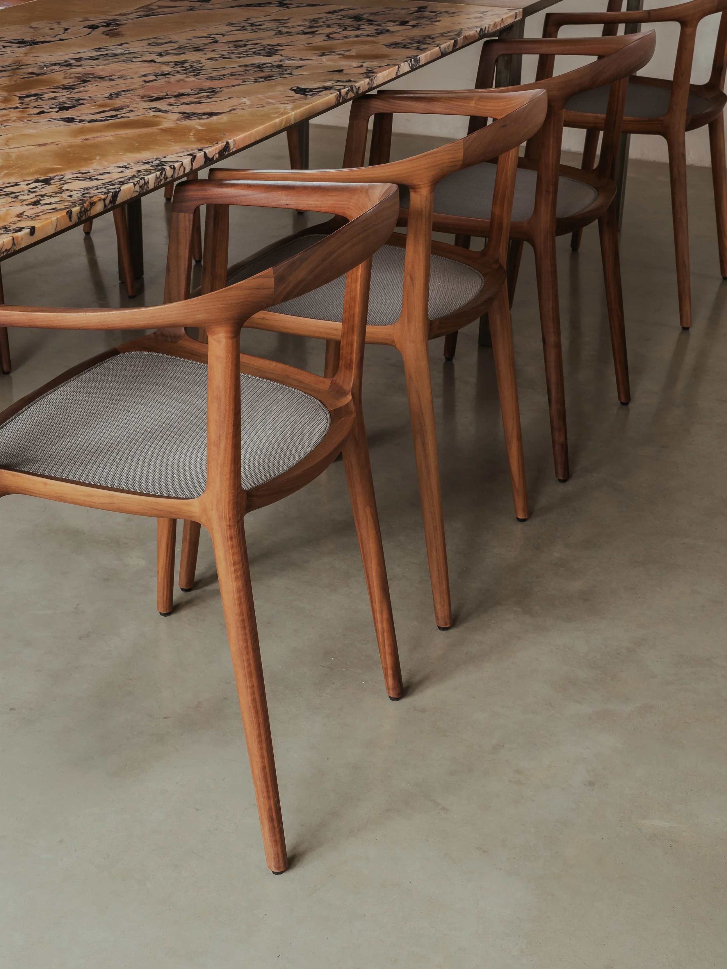 A row of wooden chairs with gray upholstered seats arranged along a marble-topped table in a modern interior space.