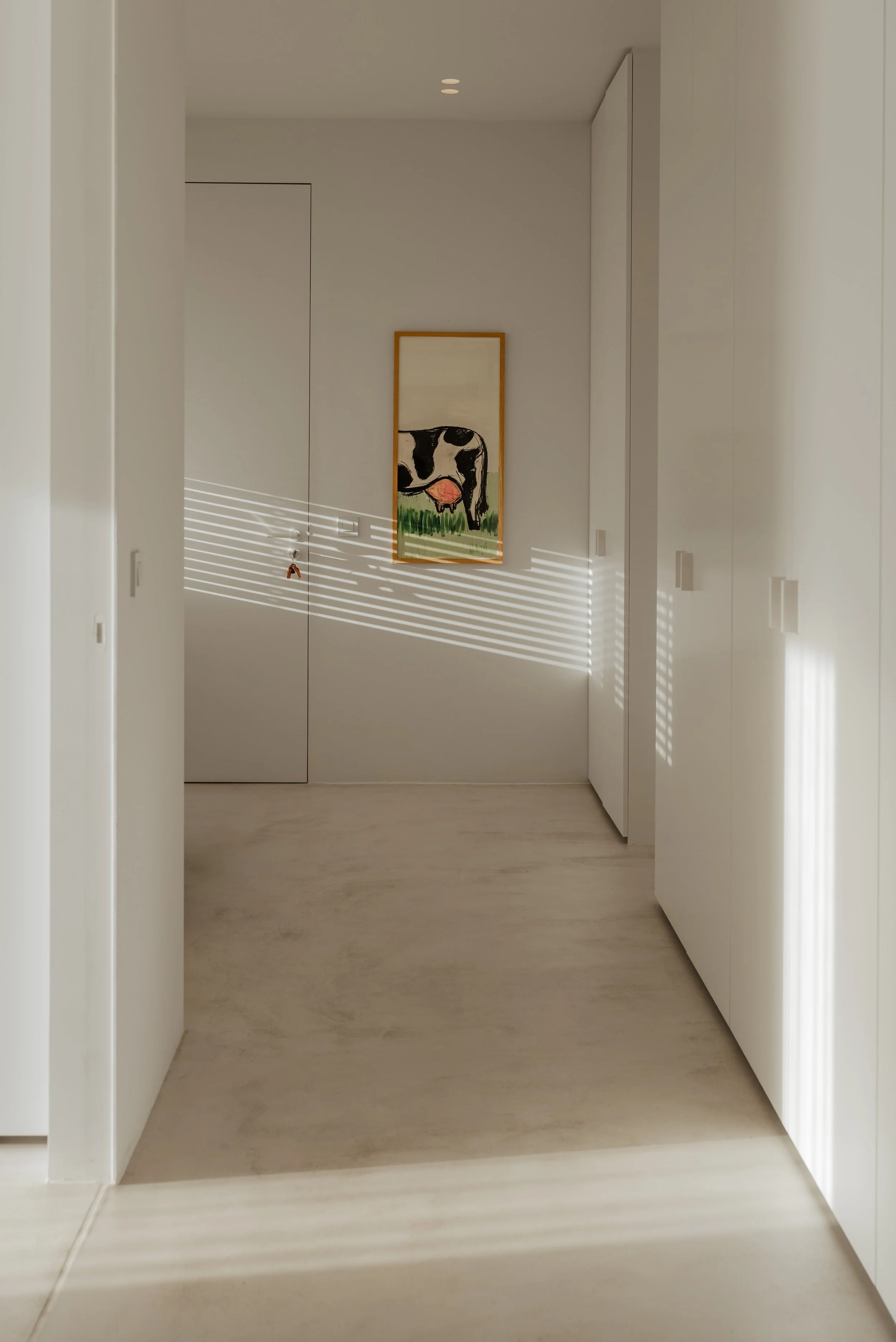 Minimalist hallway with white walls, closed white doors, a framed artwork of a cow with an udder, and sunlight creating striped shadows through blinds.
