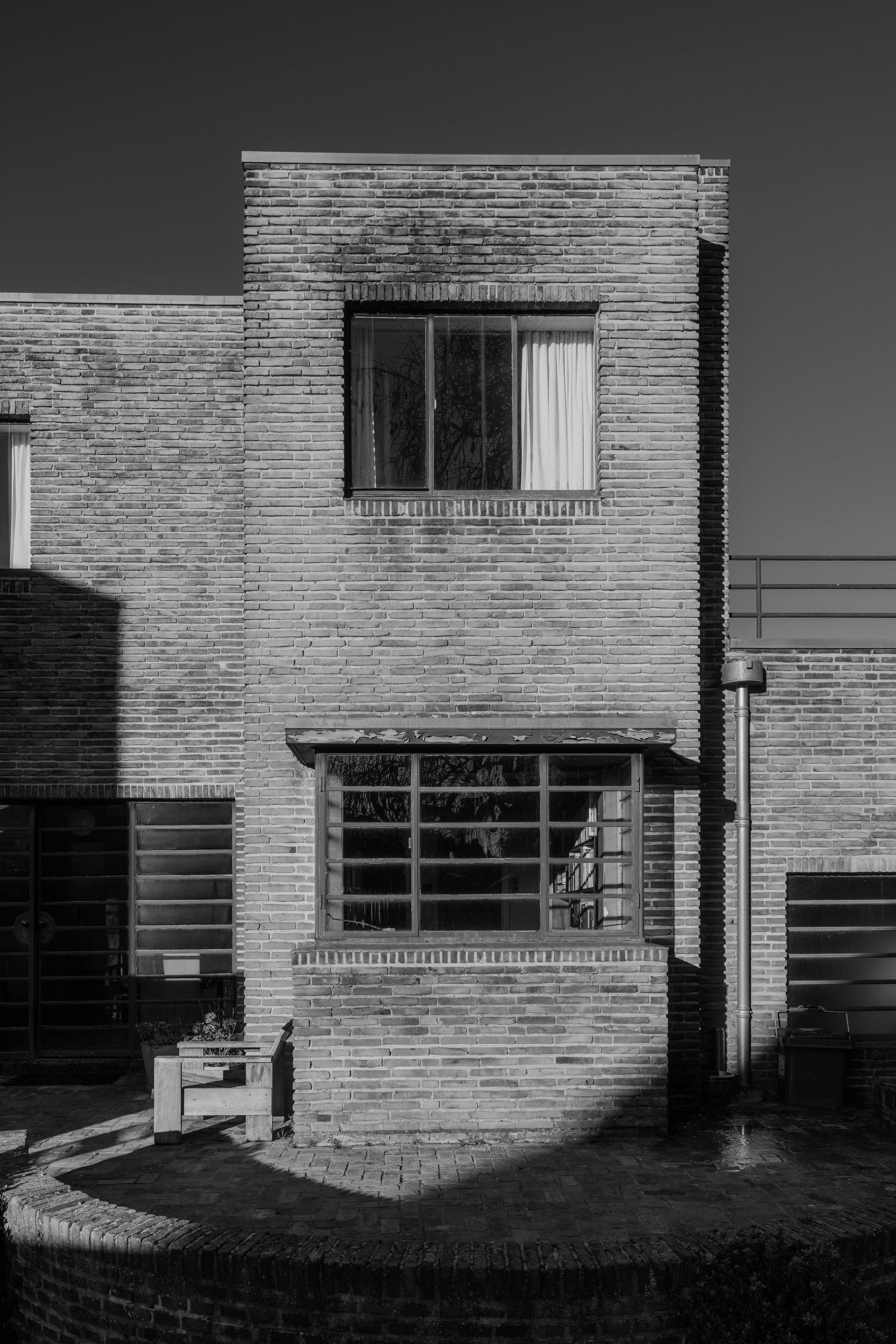 Black and white photo of a brick building with two windows, one on the upper floor and one on the lower floor. The upper window has curtains, and the lower window has multiple panes. Shadows and sunlight are visible on the building facade.