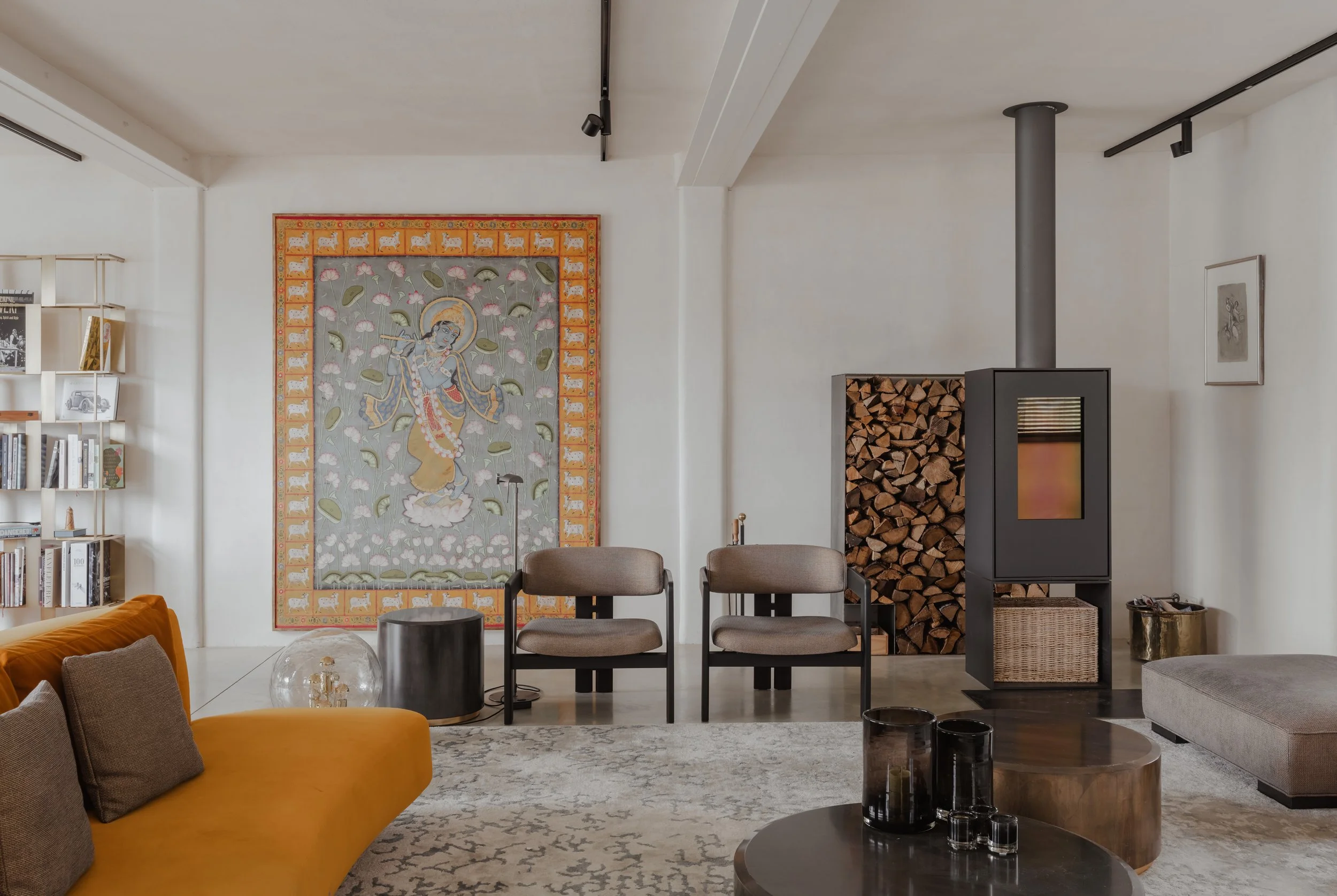 Modern living room with a mustard yellow sofa, a light-colored area rug, black chairs, a stacked firewood behind a black stove, and wall art including a large tapestry and a framed picture.