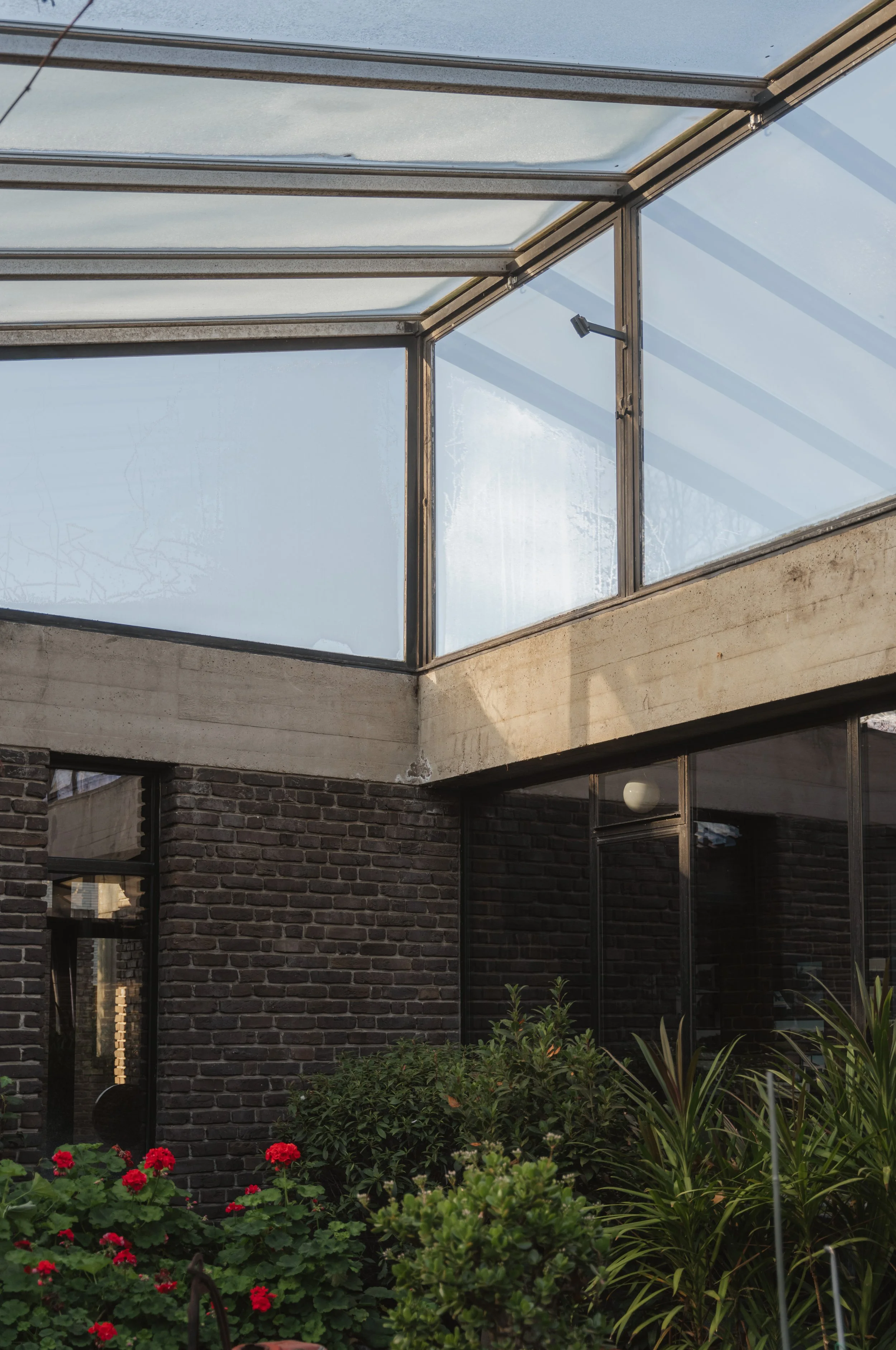 A modern building with large glass windows and a concrete overhang, featuring a garden with red flowers and various green plants.