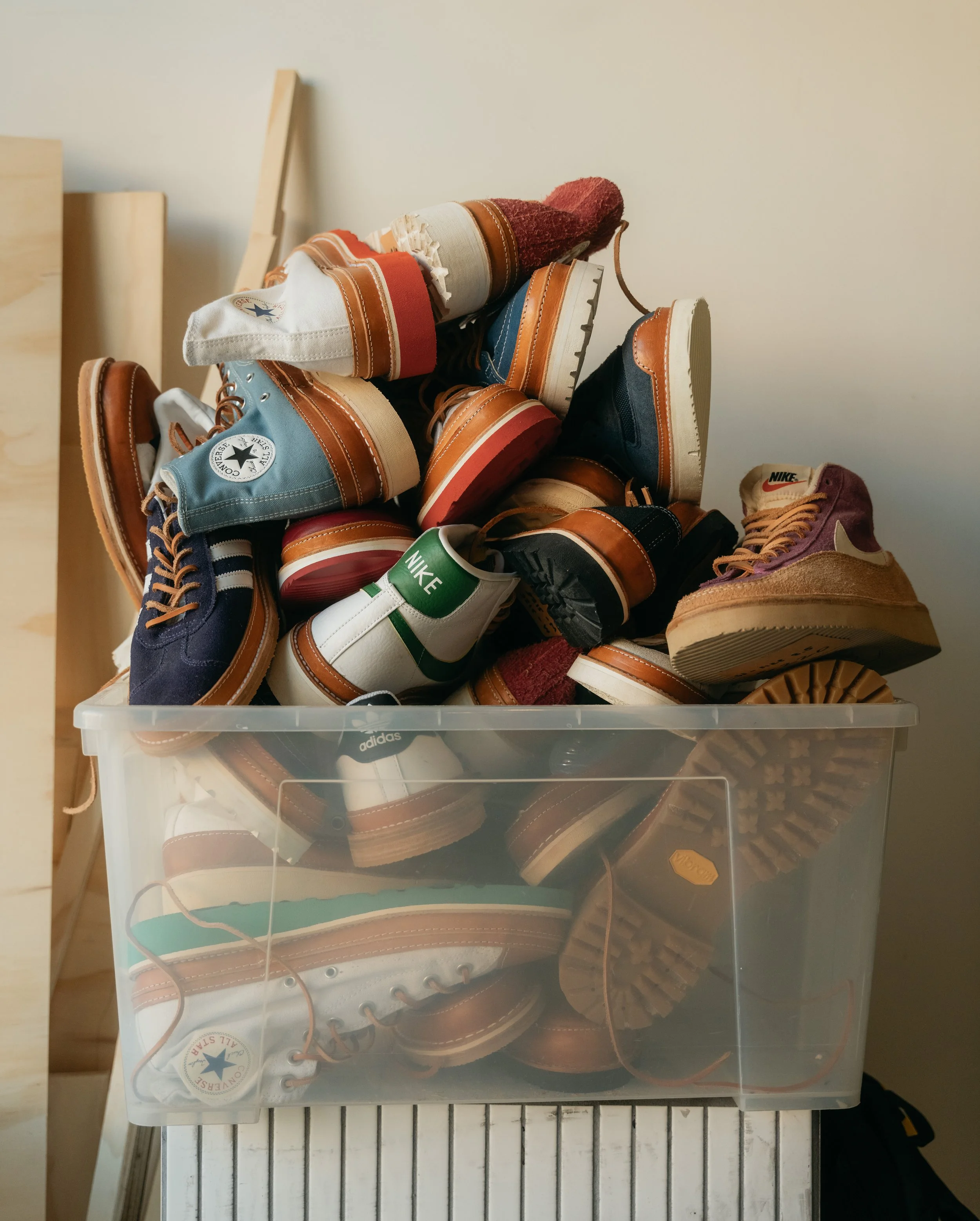 A clear plastic storage bin filled with various sneakers, some spilling out on top, in front of a plain wall.