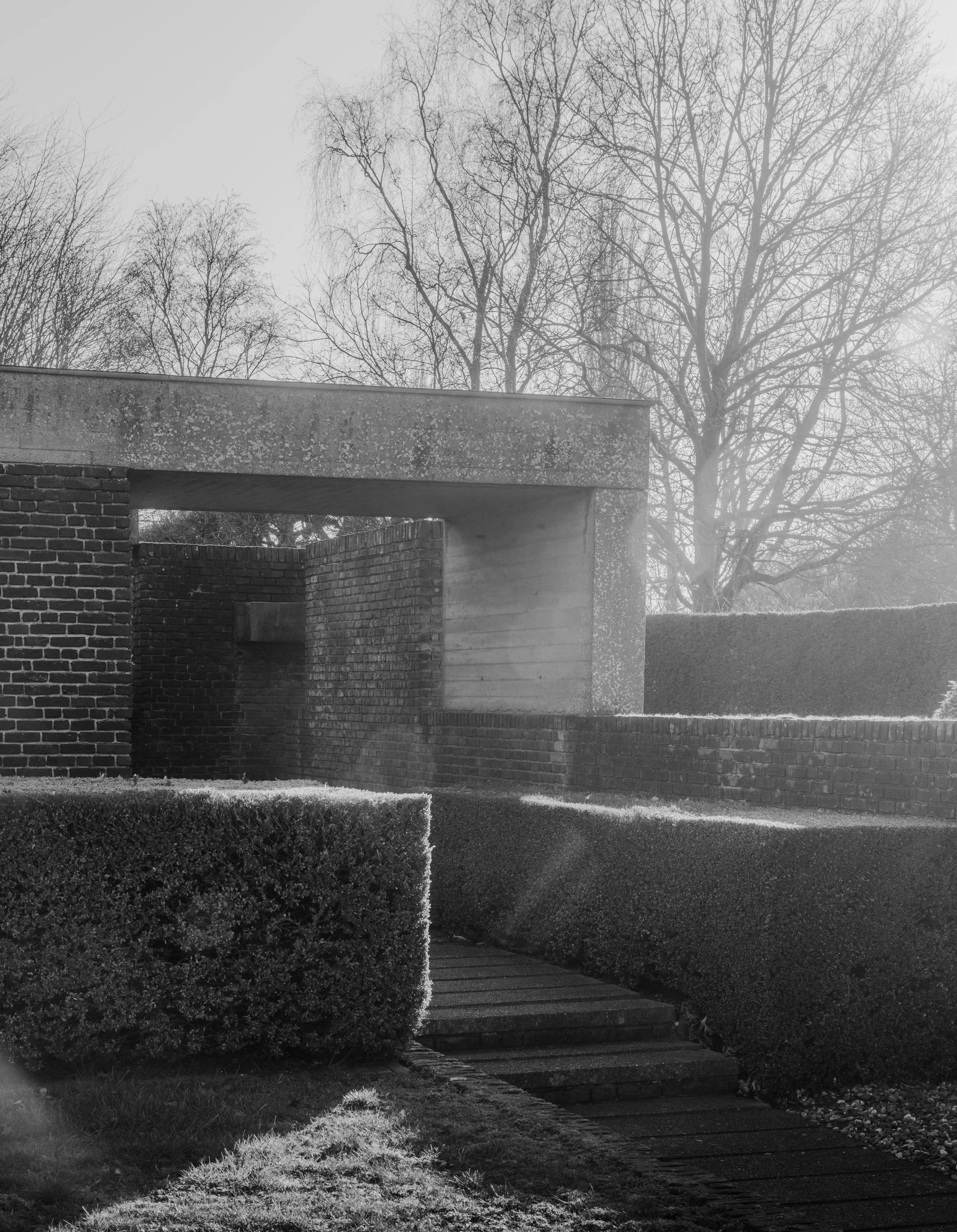 A black and white photograph of a garden pathway leading to a brick and concrete structure with trees in the background.