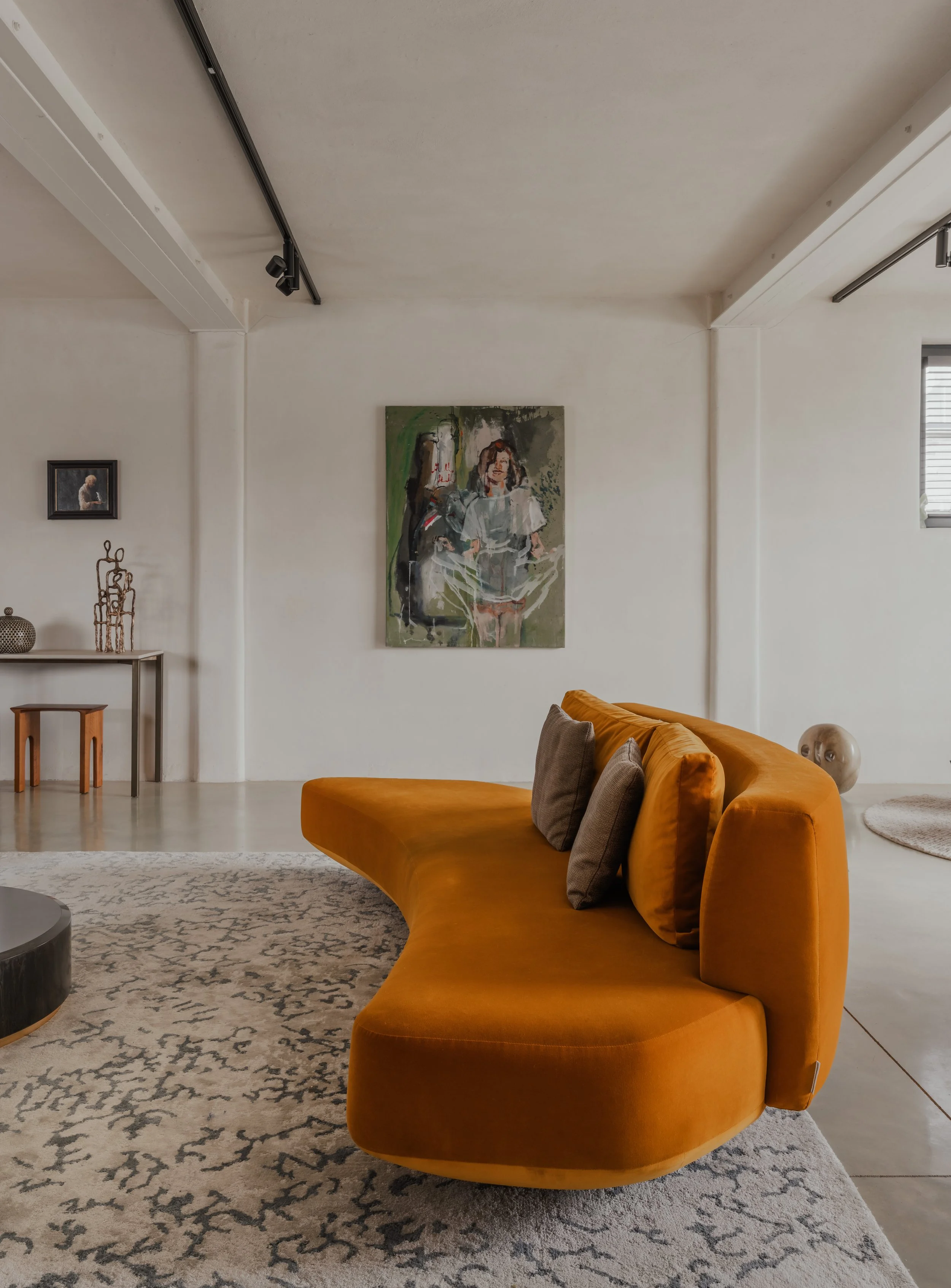 Modern living room with an orange curved sofa, gray pillows, abstract paintings on white walls, side table with decorative objects, and large windows letting in natural light.