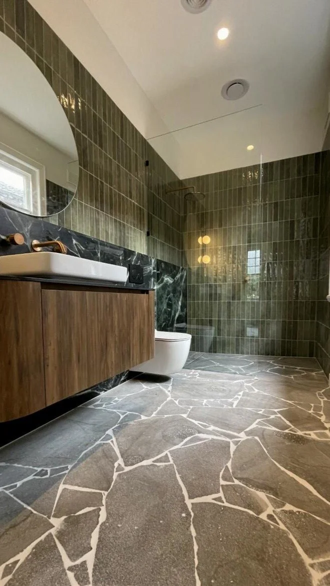 Modern bathroom with green tiled shower, white toilet, black marble vanity with a white sink, and a stone patterned floor.