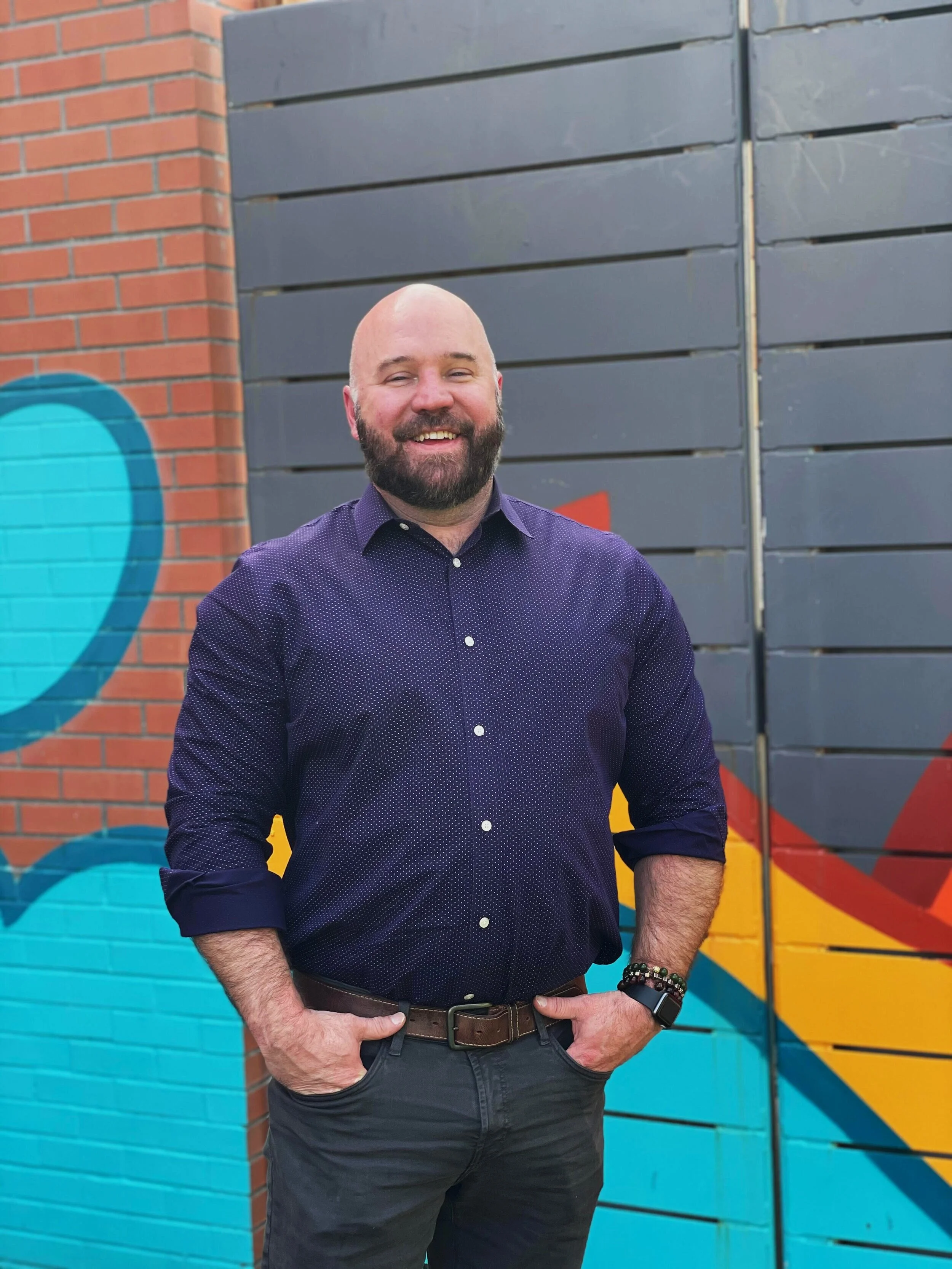 Therapist Shawn Chrisman smiling outdoors in front of a colorful mural, offering a warm and welcoming presence.
