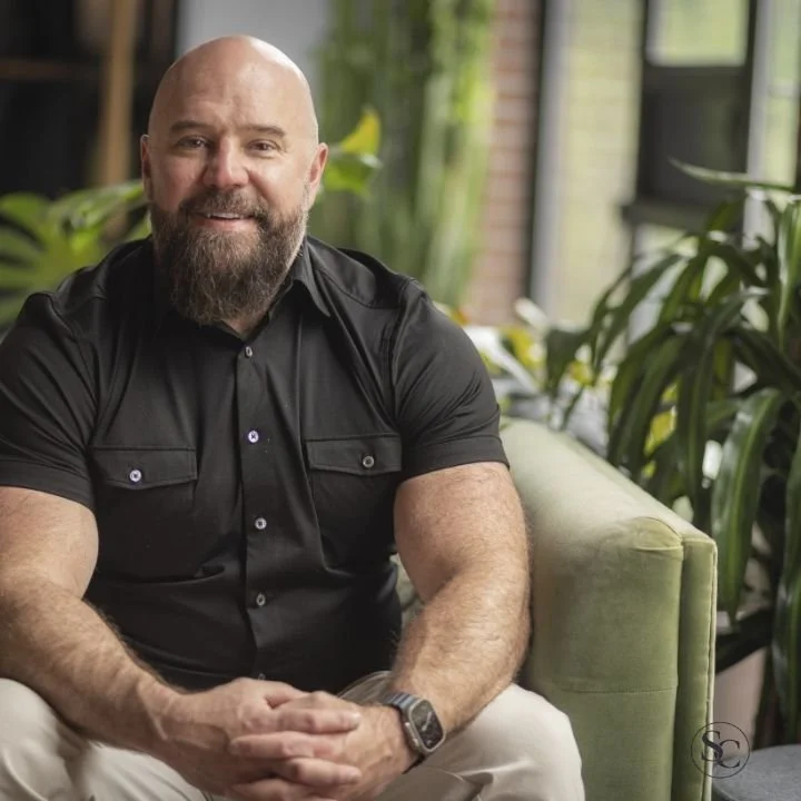 Therapist sits on a green sofa in a modern Dallas therapy office with plants and natural light behind him.