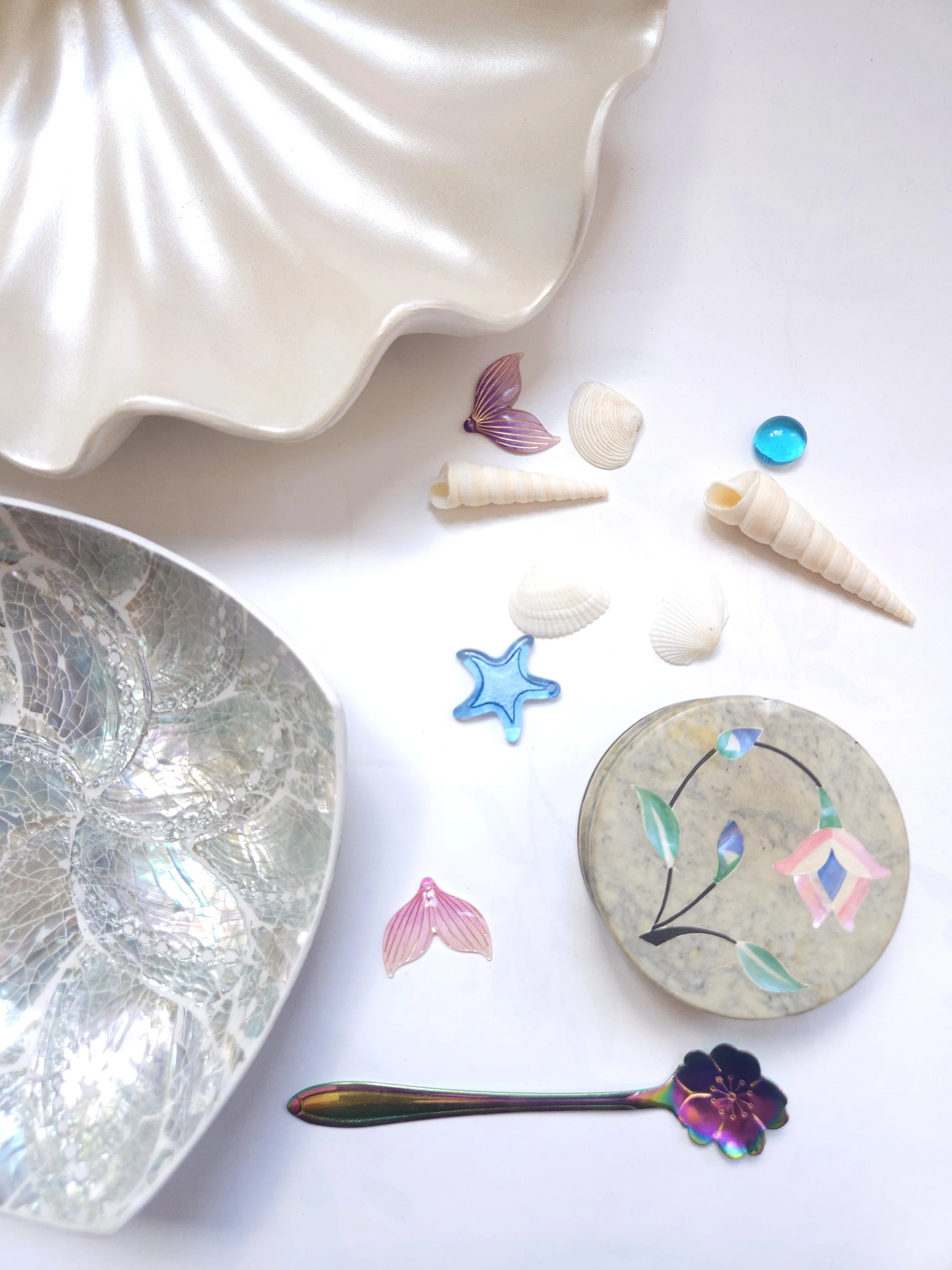 Decorative collection of seashells and colorful glass ornaments on a white surface, including a large shell, a small shell, a conch shell, and various sea-themed glass pieces in different shapes and colors.