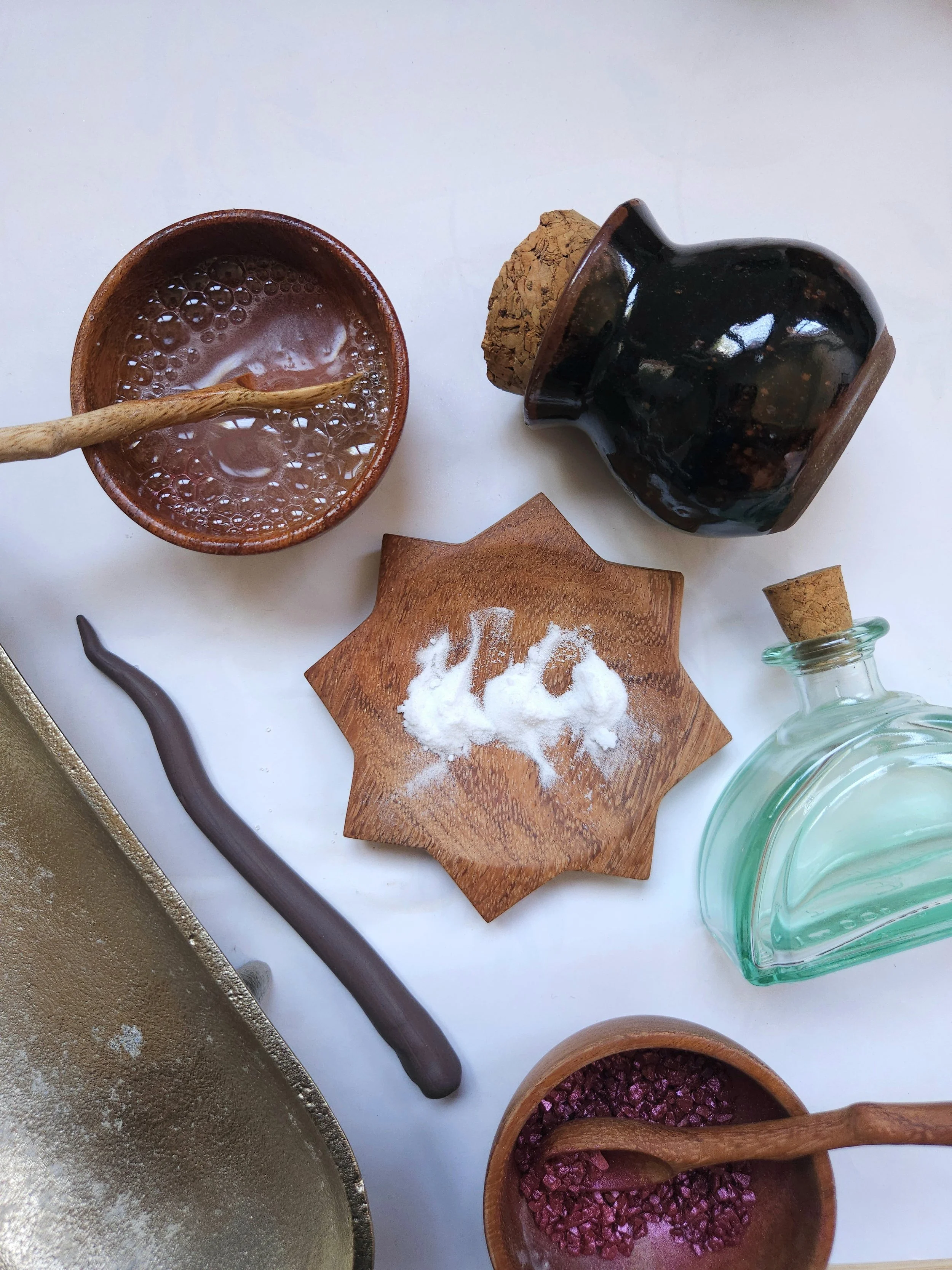 Various jars and bowls with different ingredients, including a wooden spoon in a small bowl, a corked glass bottle, a wooden dish with white powder, and a bowl with pinkish salt or crystals, arranged on a white surface.