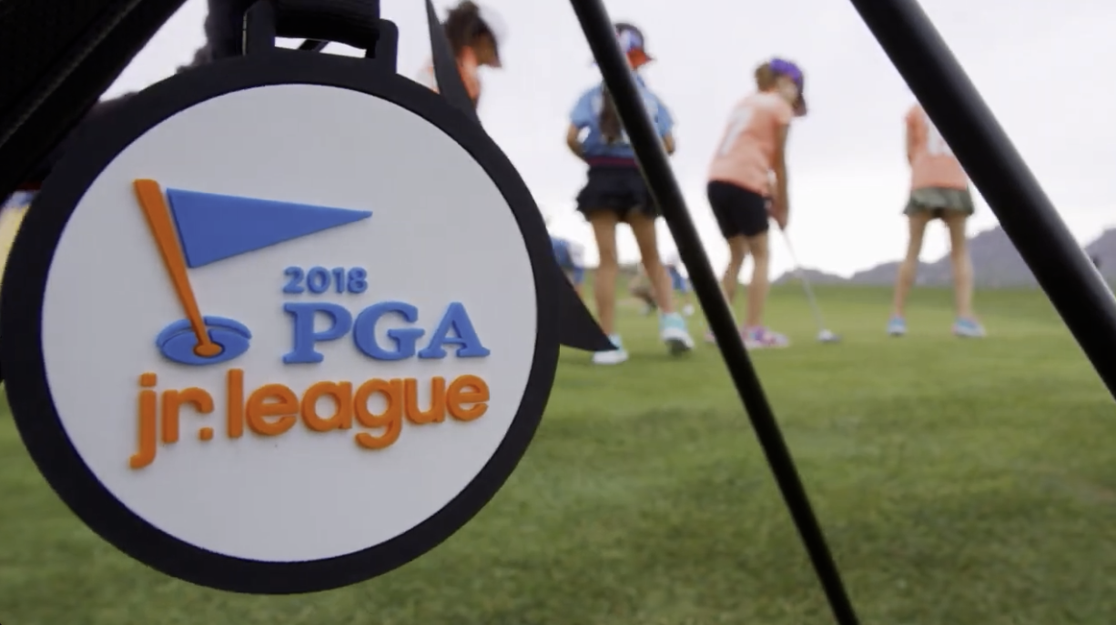 Close-up of a medallion with the 2018 PGA Jr. League logo, featuring a golf flag and hole, with young children playing golf in the background on a golf course.