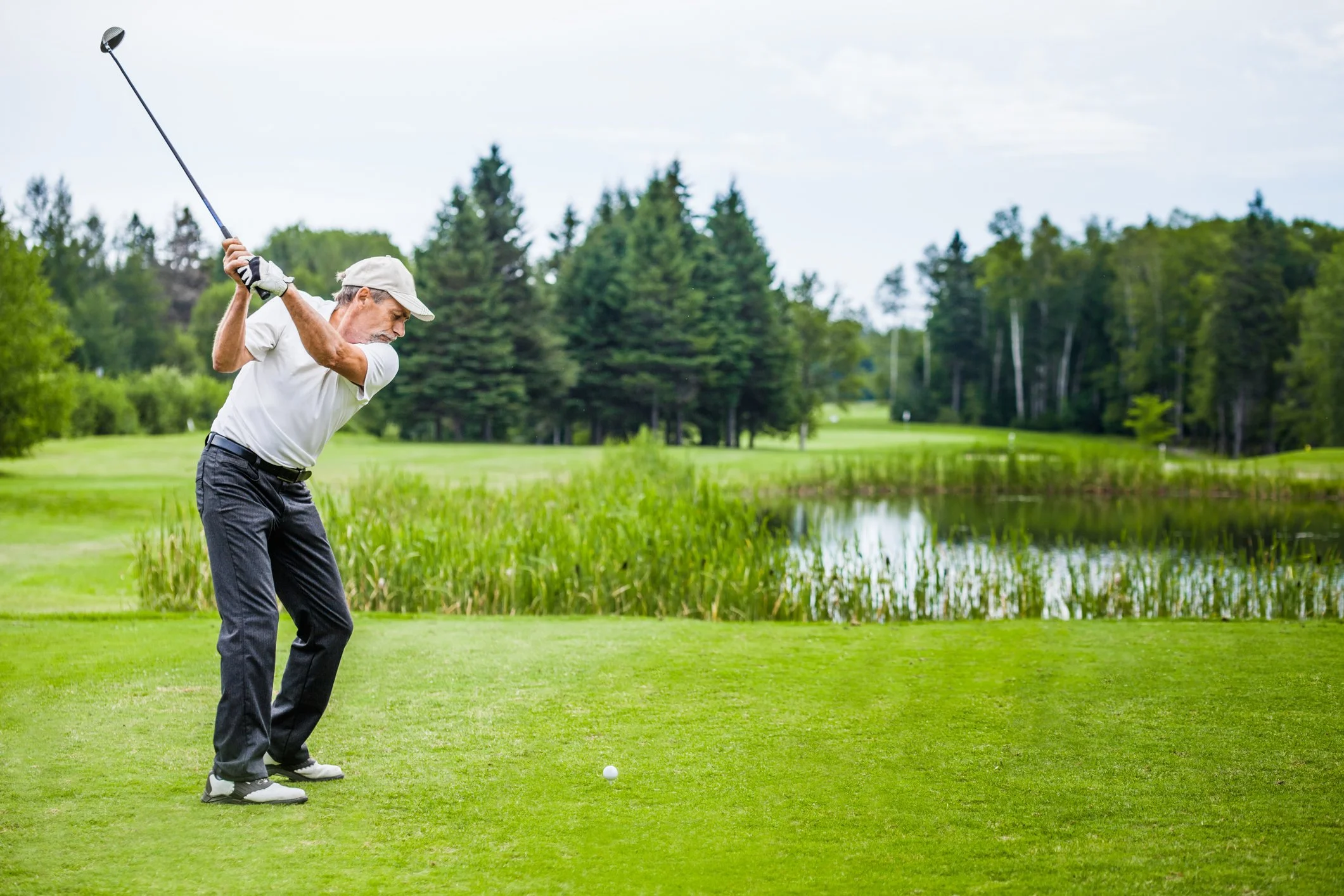 An elderly man playing golf on a lush course next to a pond, preparing to swing his golf club.