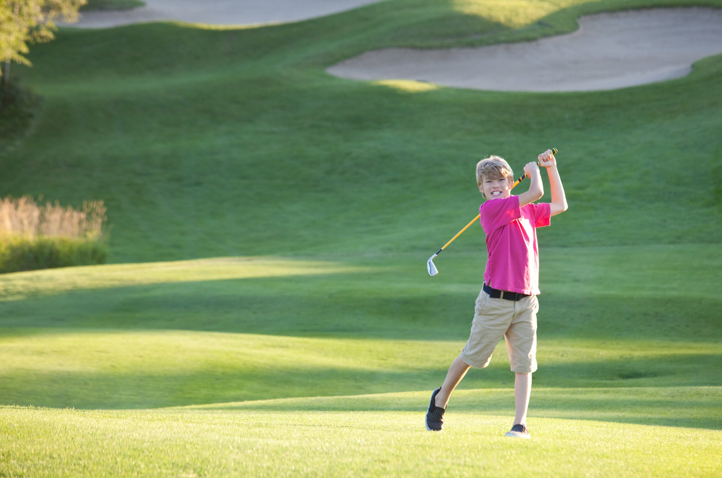 A young boy wearing a pink shirt and khaki shorts is swinging a golf club on a golf course during daytime.