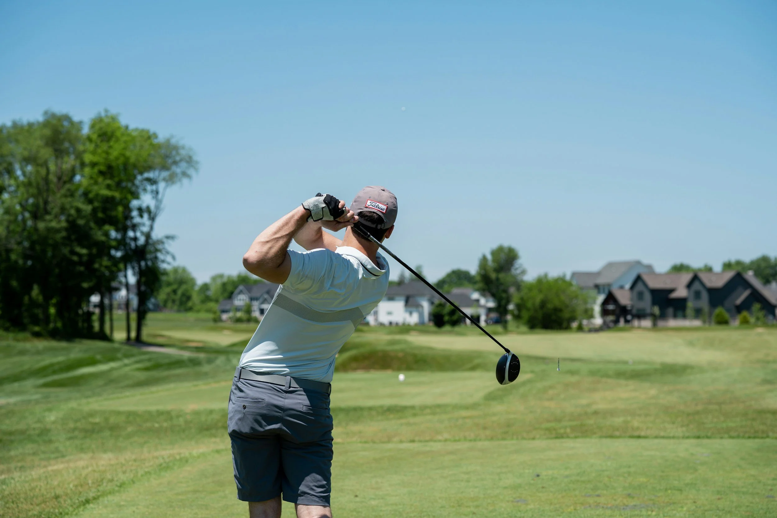A man in a white shirt and gray shorts playing golf on a sunny day, swinging a driver club on a golf course with houses in the background.
